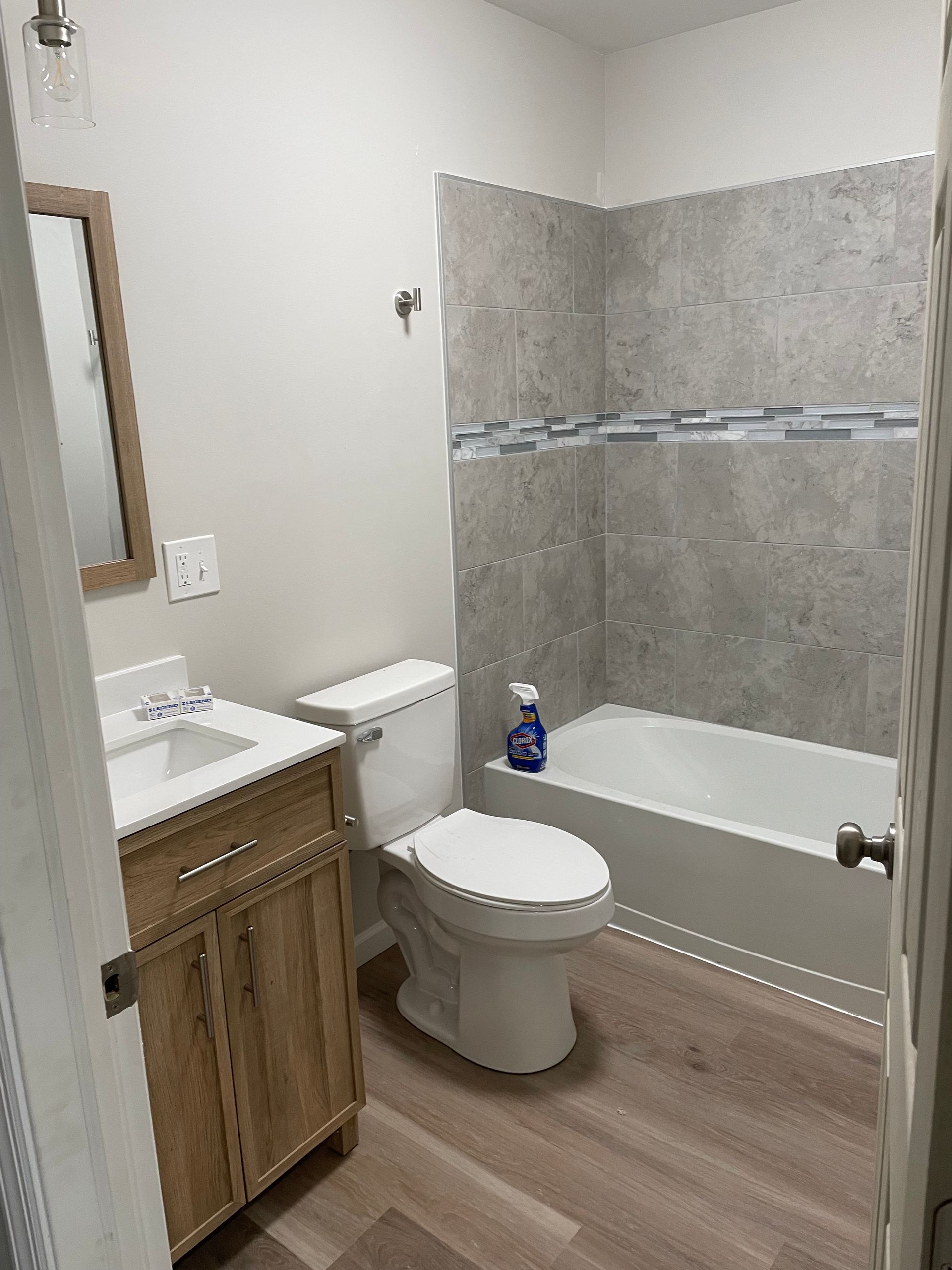 A small bathroom with light wood cabinets, a white vanity sink, a toilet, and a bathtub with gray tile surround.