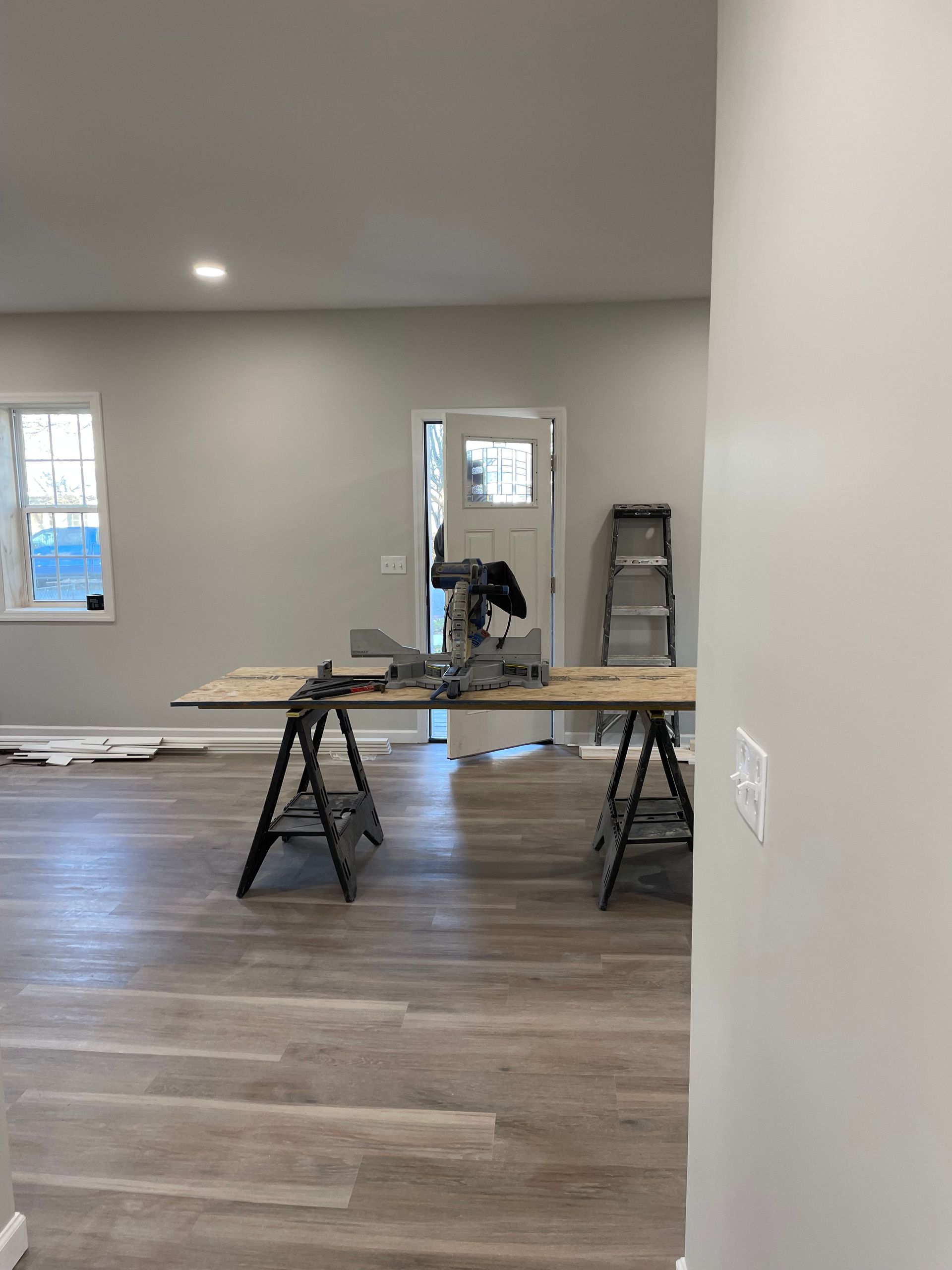 A miter saw sits on a plywood table supported by sawhorses in an unfurnished room with gray walls and wood flooring.