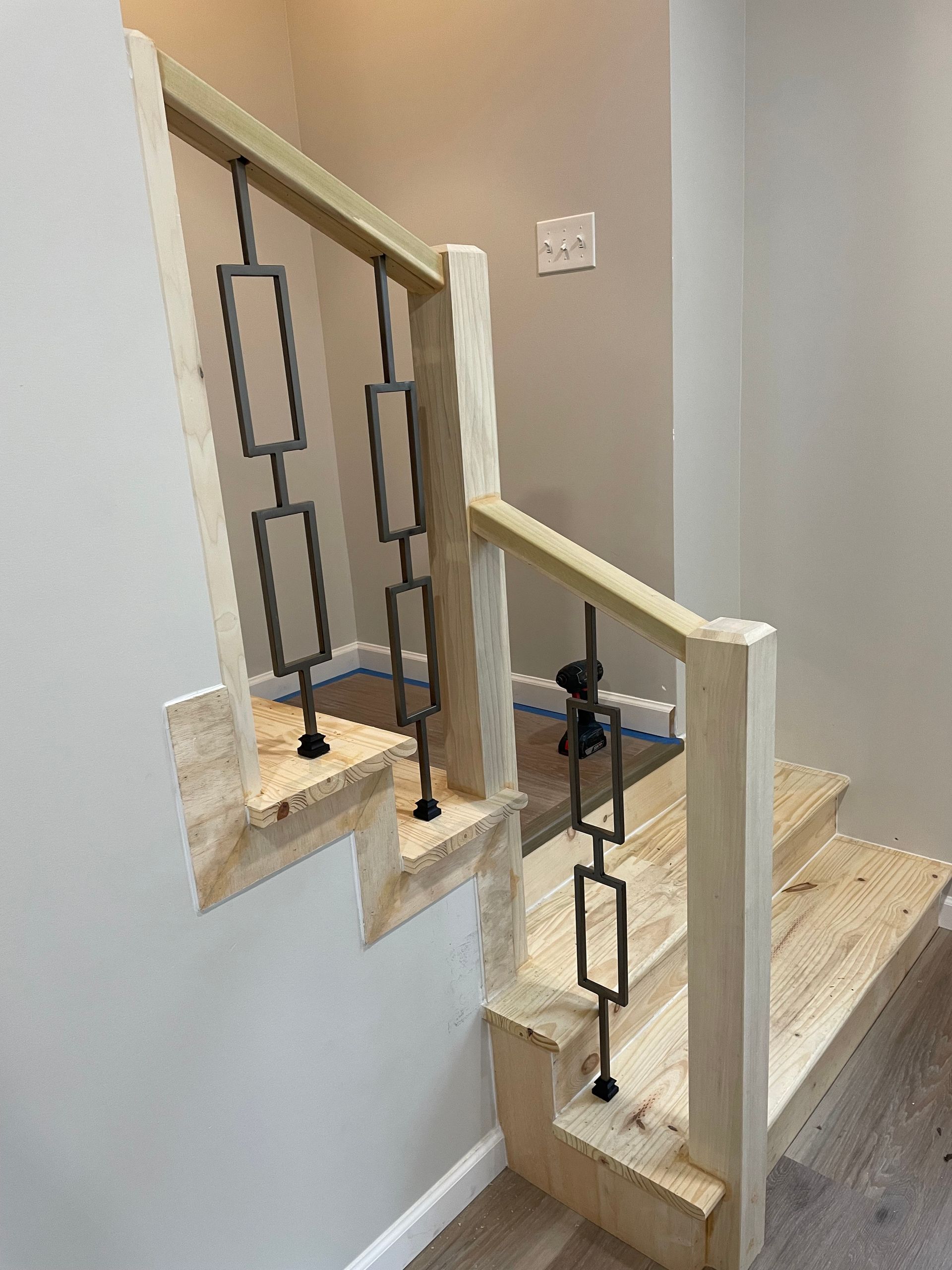A light wood staircase under construction, featuring vertical black metal railings with geometric rectangular accents.