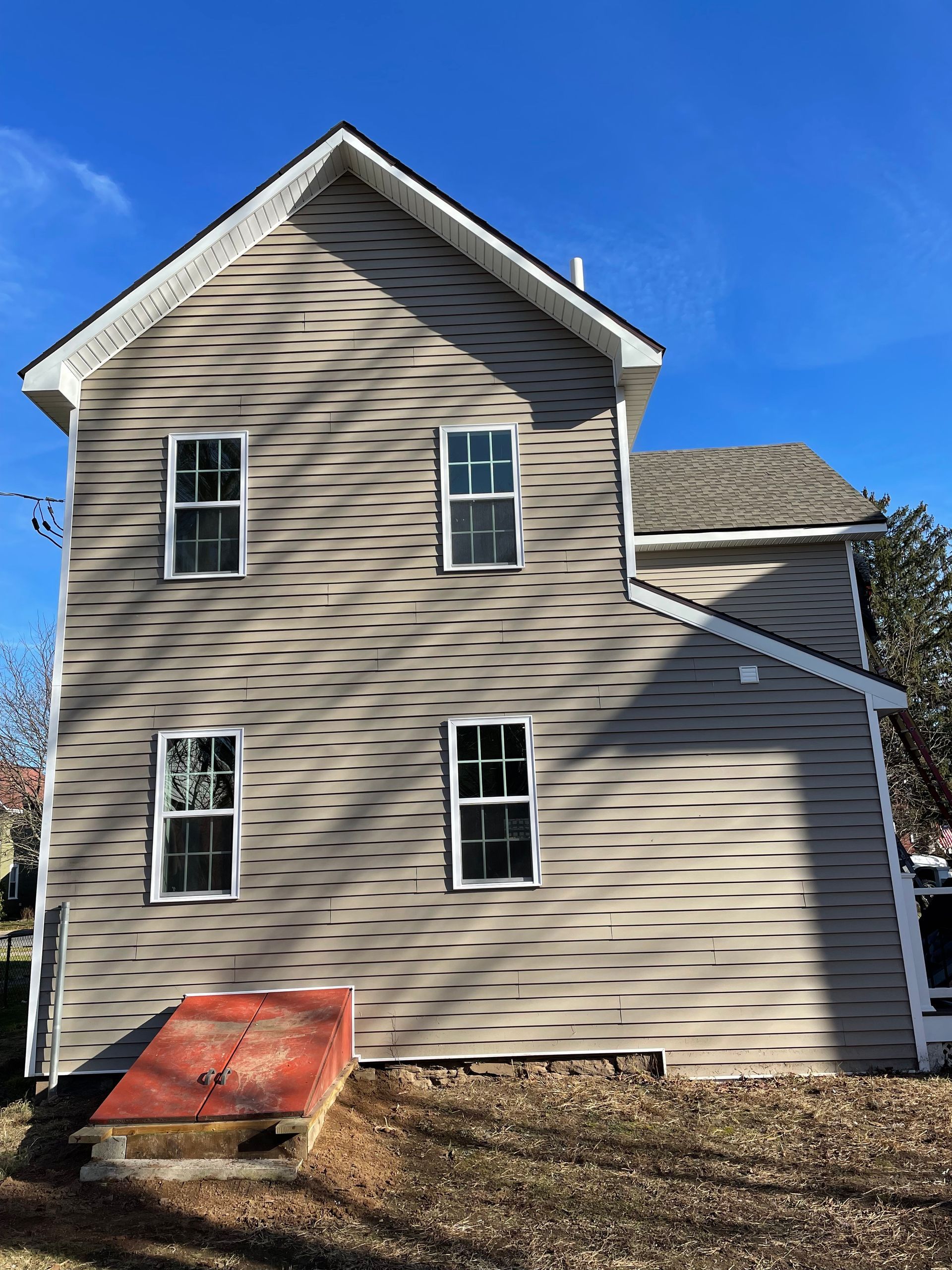 A two-story tan house exterior under a clear blue sky, featuring a red cellar door and wavy tree shadows on the siding.