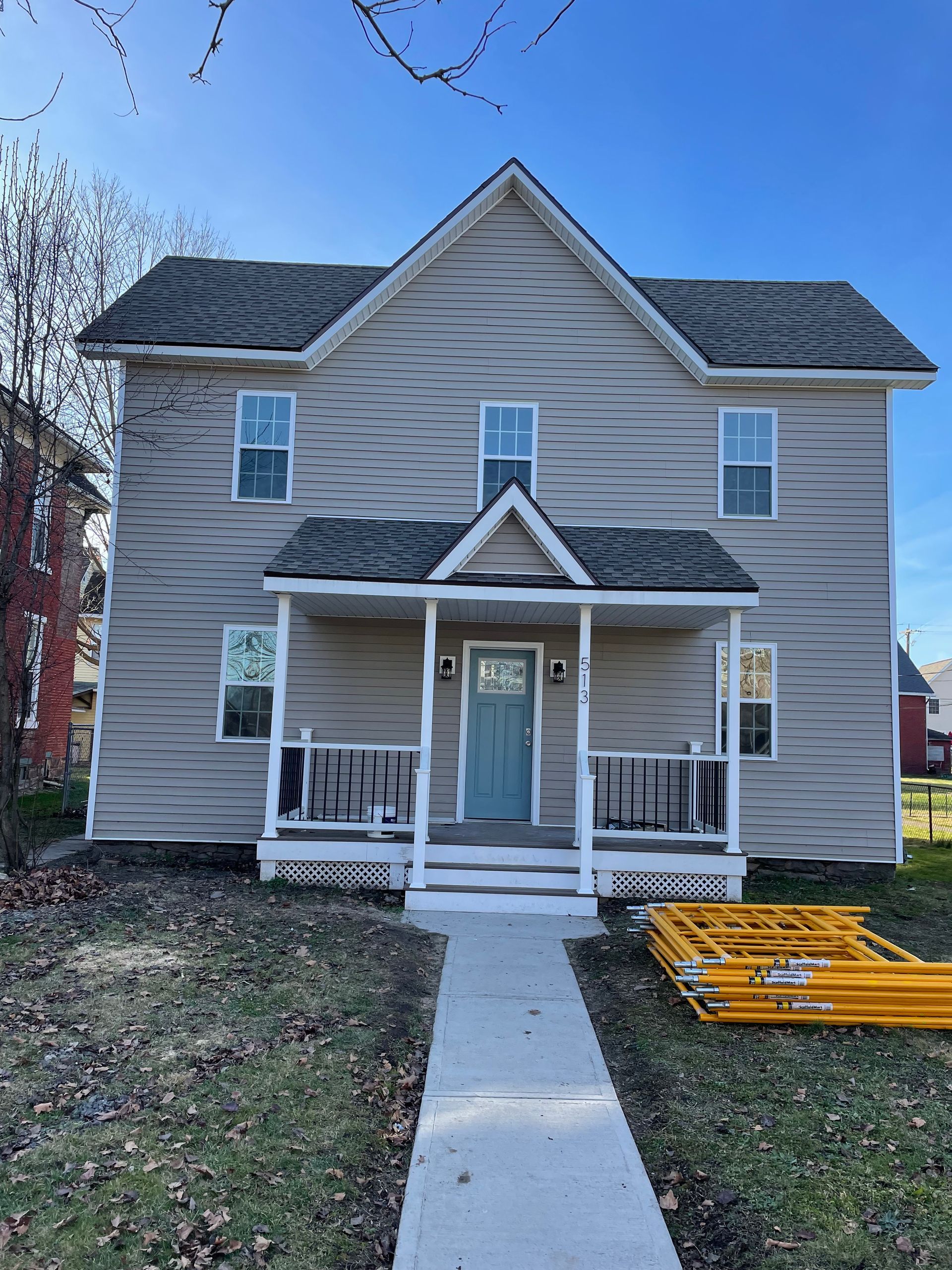 A two-story tan house with a covered front porch, teal front door, and gray roof, viewed from a sidewalk on a sunny day.
