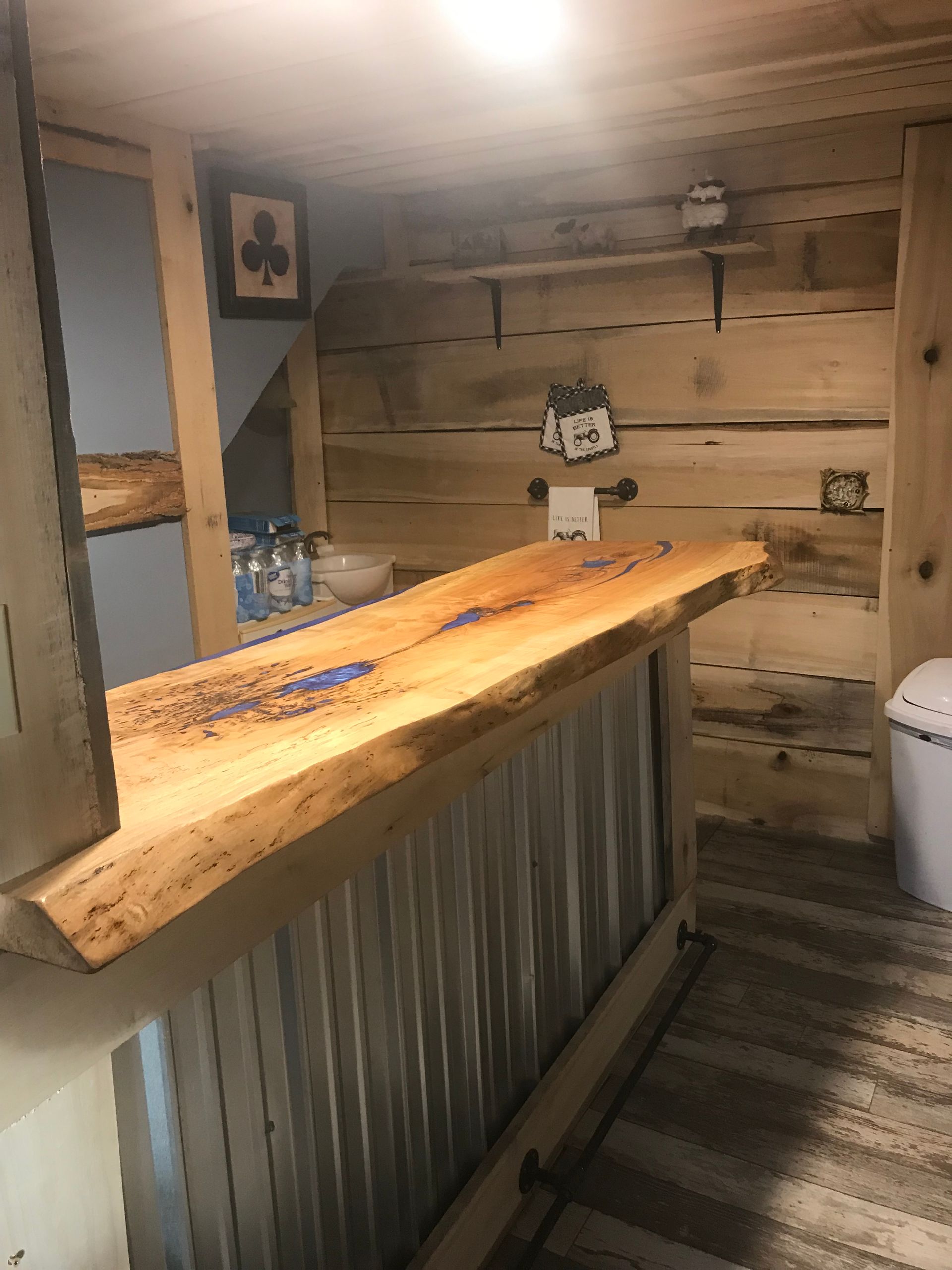 A rustic home bar with a live-edge wooden countertop, corrugated metal base, and log-style paneled walls.