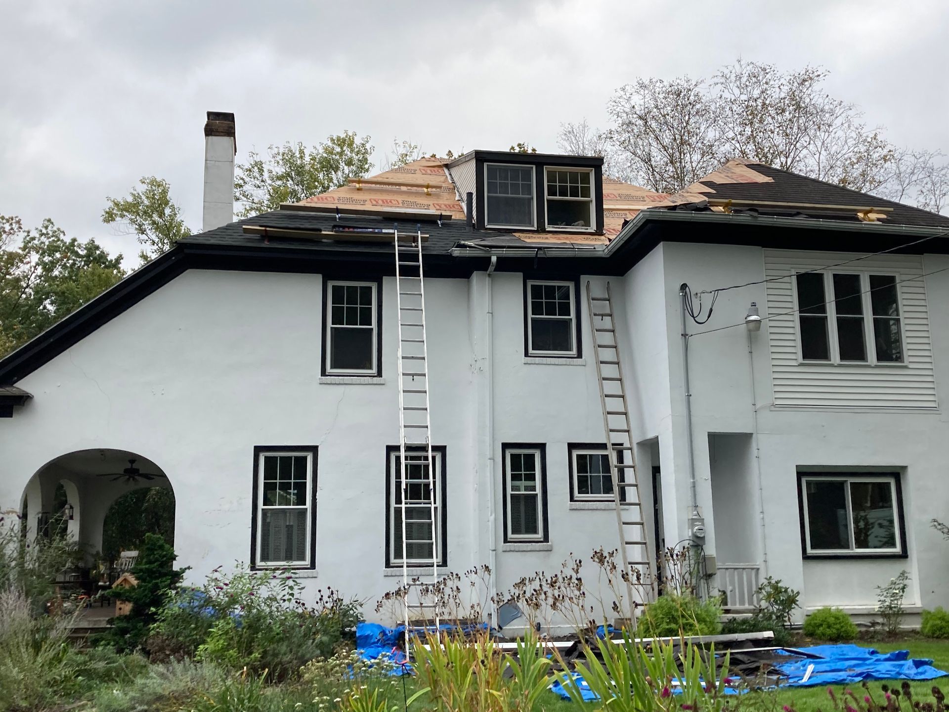 A white house with black trim undergoing roof repairs, featuring two ladders leaning against the exterior walls.