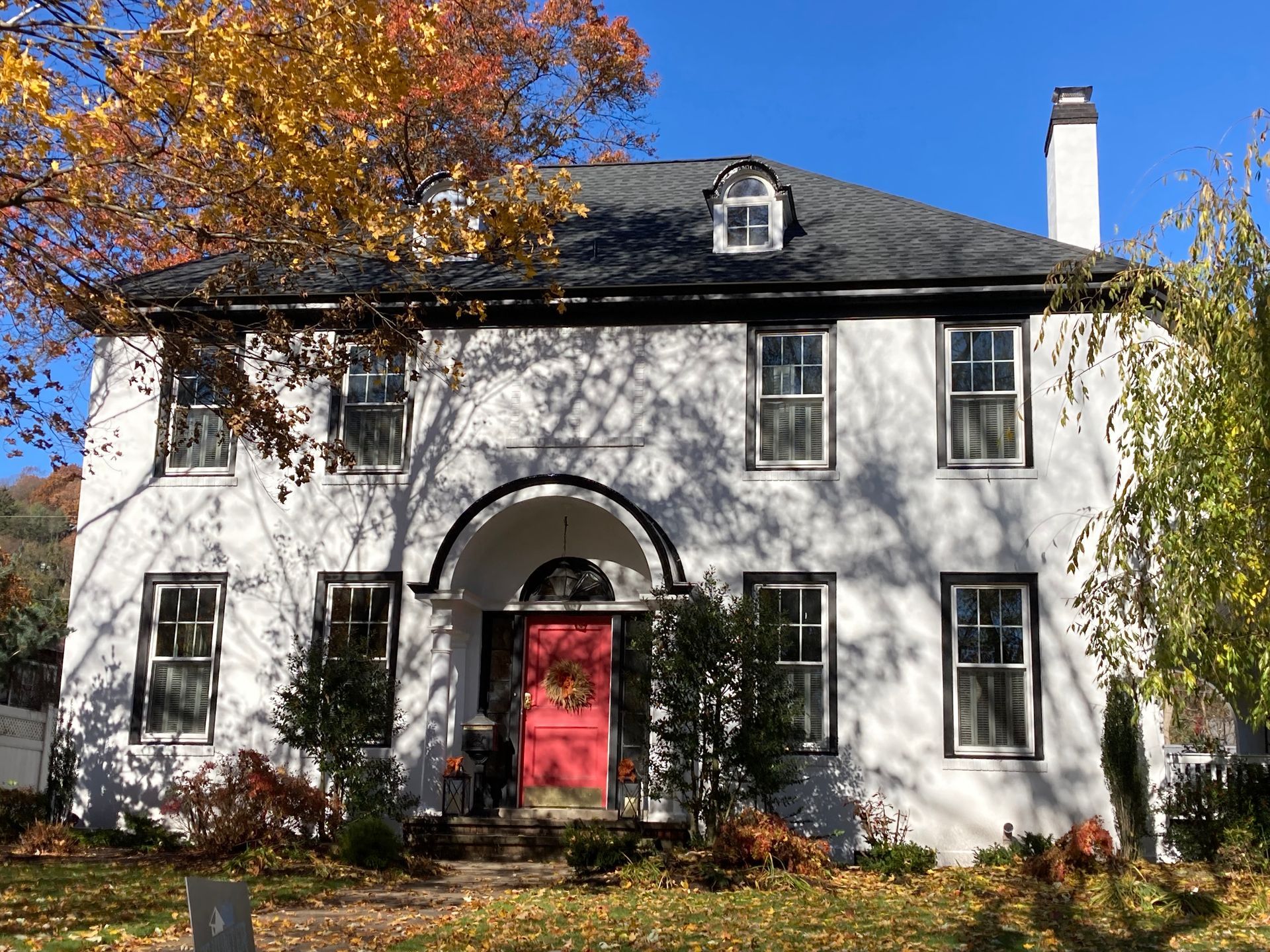 Two-story white house with a red front door, black trim, and two dormer windows under a blue sky on a fall day.