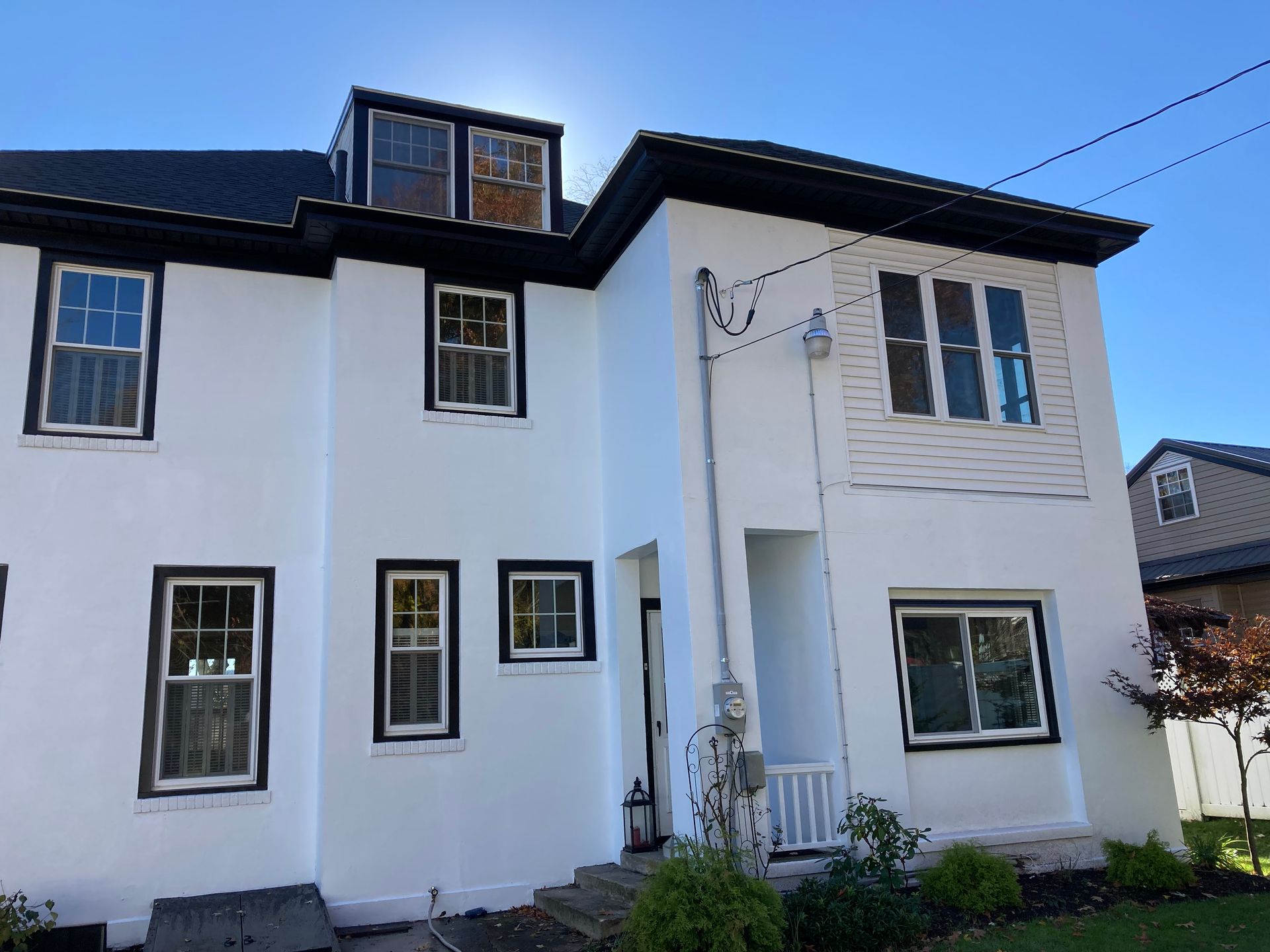 A white, two-story house exterior under a clear blue sky, featuring dark-framed windows and a small attic dormer.