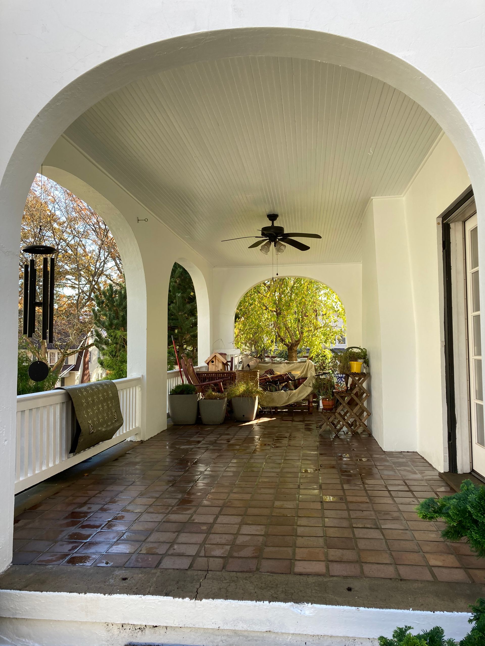 A covered arched porch with white walls, terracotta tile flooring, a ceiling fan, and outdoor seating.