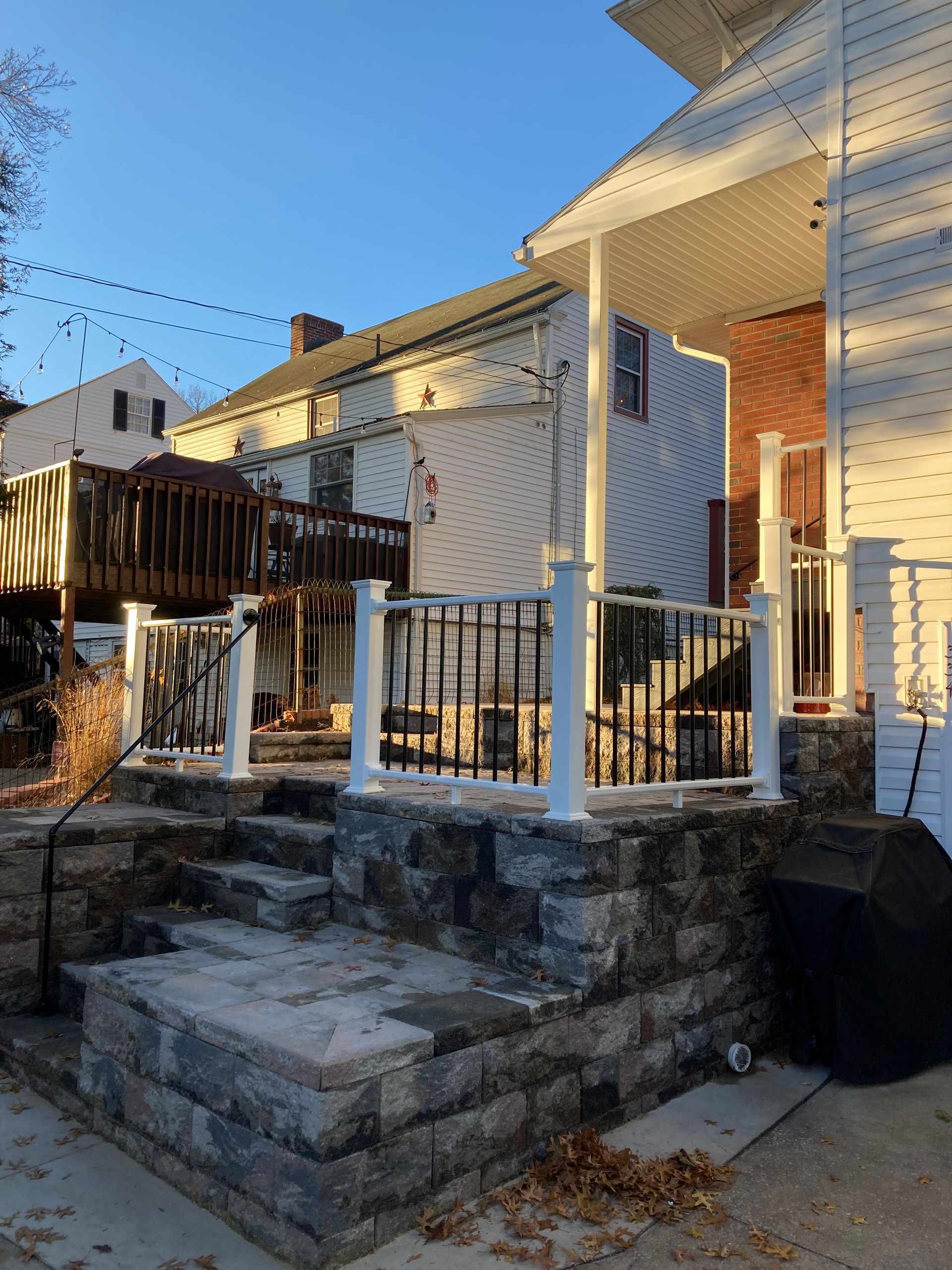 A stone patio with white railings and stairs, connected to a white house with a wooden deck during sunset.