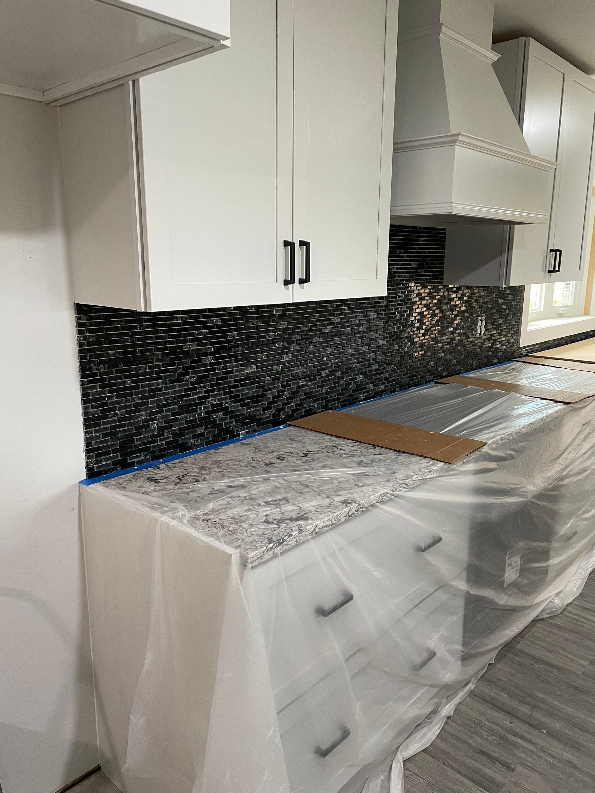 A kitchen with white cabinets, a black tiled backsplash, and granite countertops covered in protective plastic.