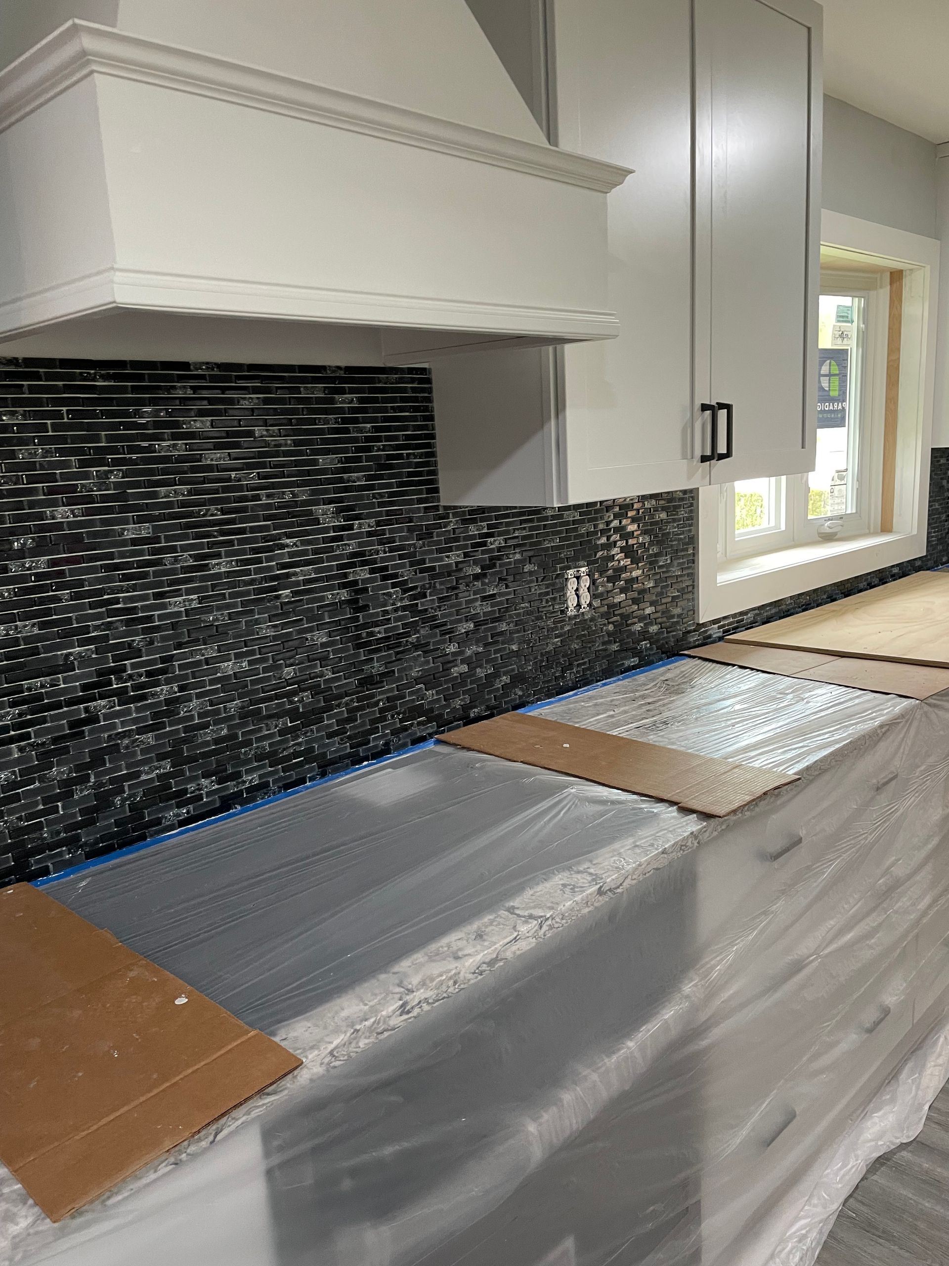 A kitchen backsplash with black textured tiles installed under a white range hood and cabinets, with covered countertops.