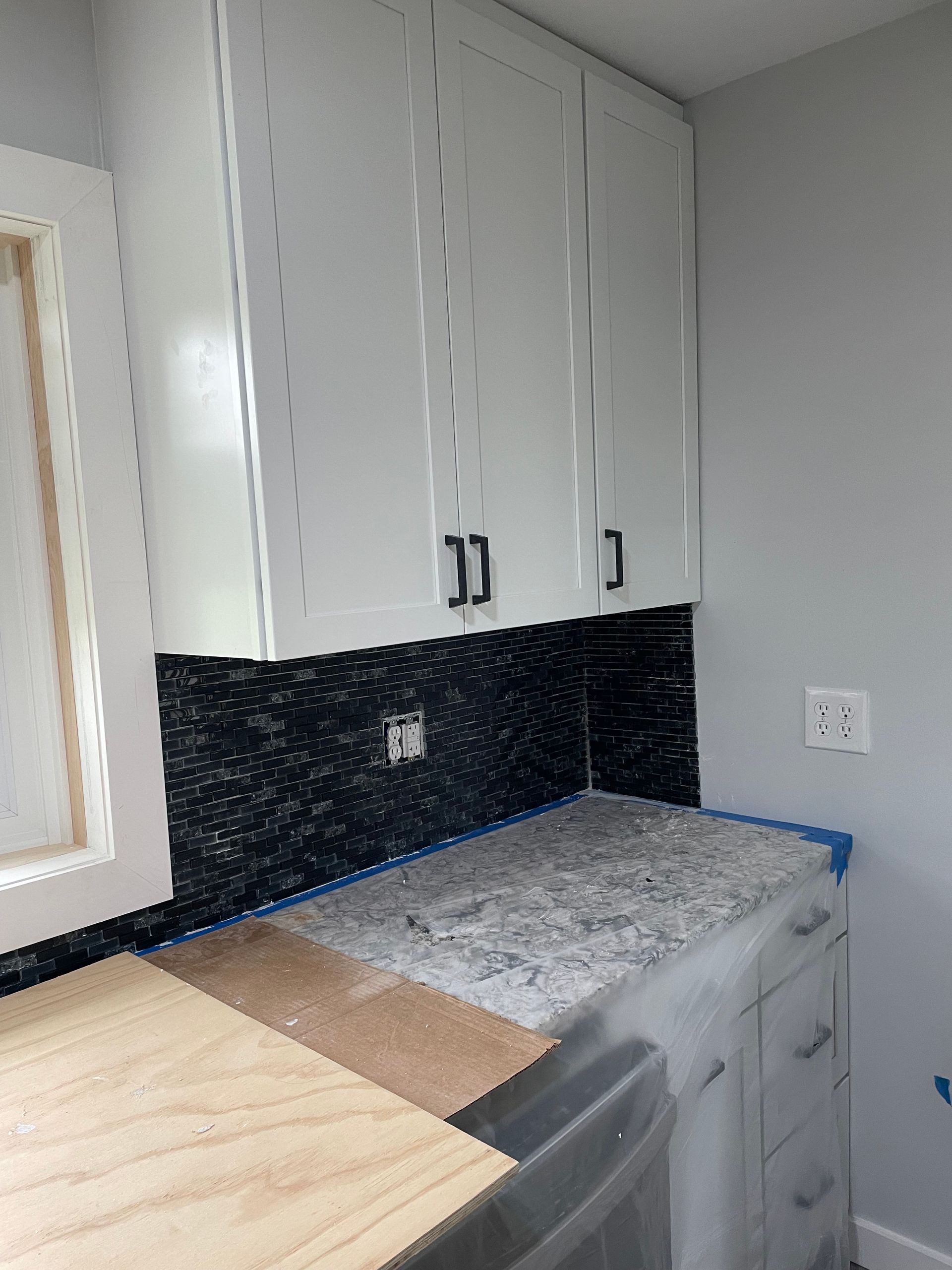 A kitchen under renovation with white cabinets, a black tiled backsplash, and a light-colored countertop.