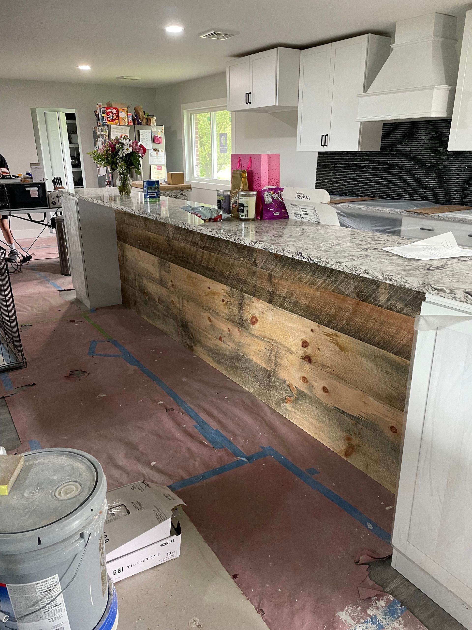 A kitchen island with a rustic wood front and granite countertop, surrounded by floor coverings during a renovation.