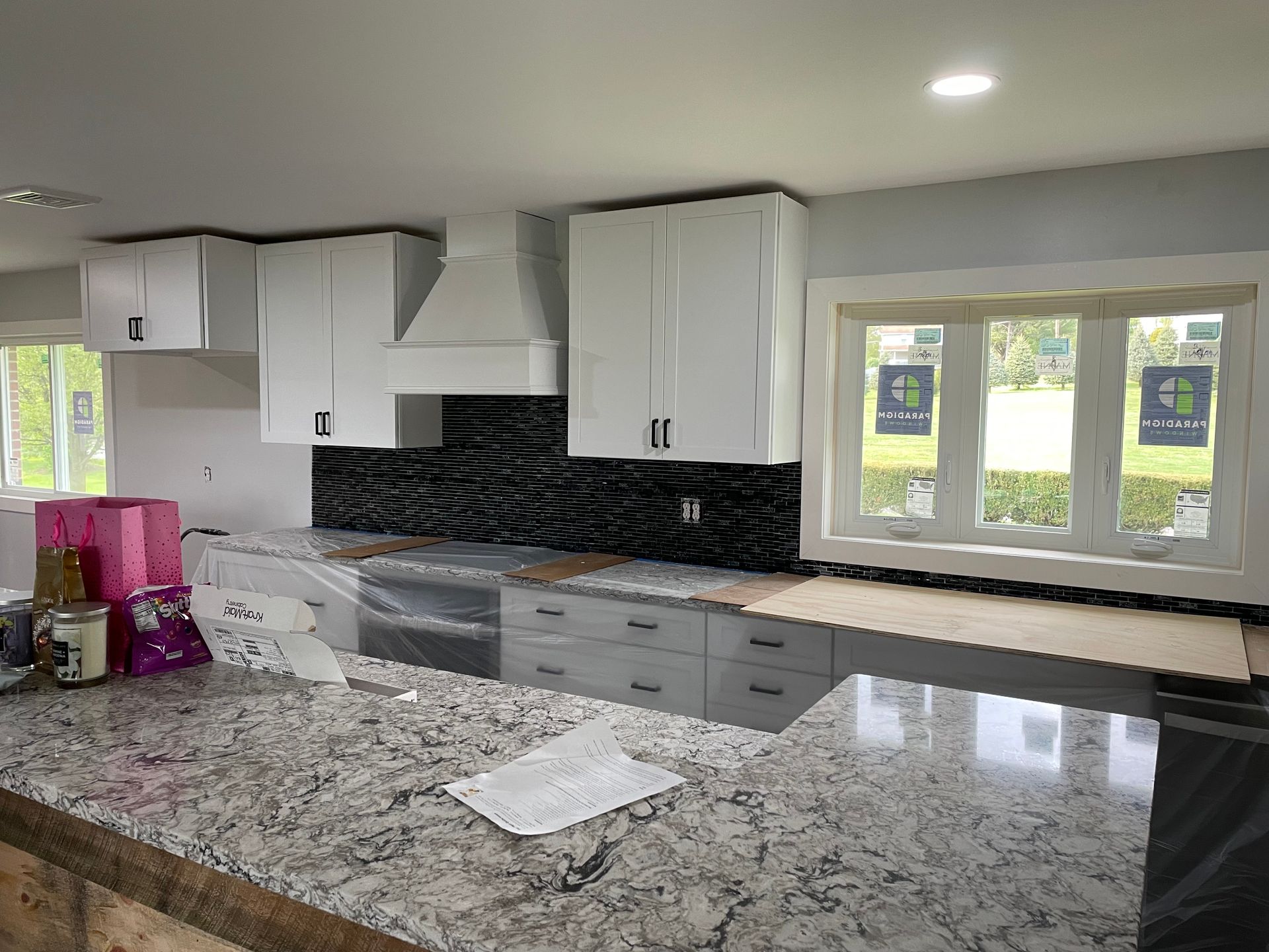 A kitchen with white cabinets, a black tiled backsplash, gray lower drawers, and a granite-topped island under construction.