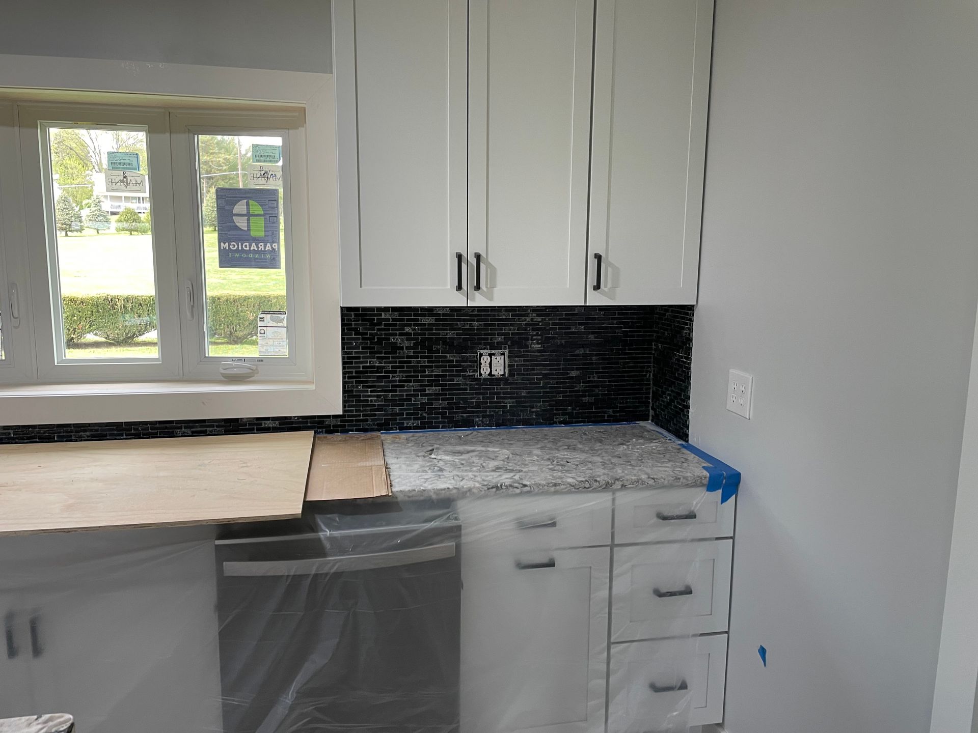 A kitchen under renovation featuring new white cabinets, a granite countertop, and a dark tiled backsplash by a window.