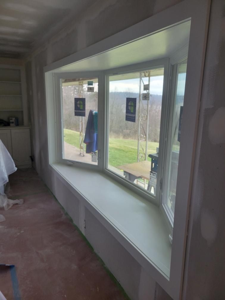 A newly installed white-framed bay window overlooks a grassy yard in a room with unfinished drywall.