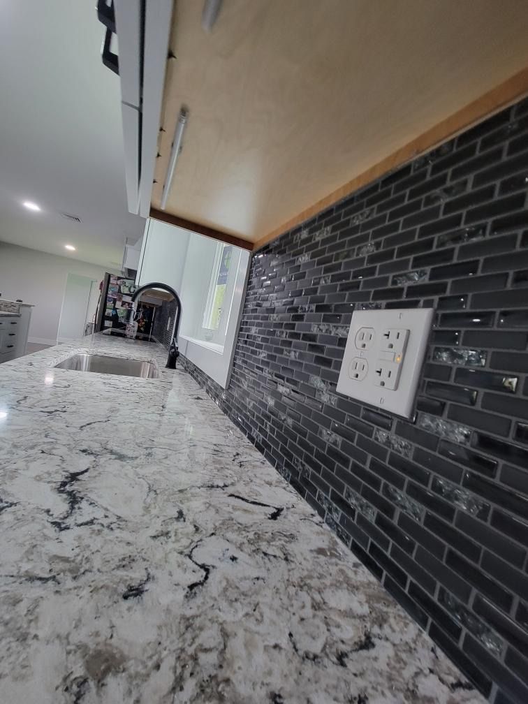 A kitchen counter with white marbled granite, a dark glass brick backsplash, and a white power outlet on the wall.