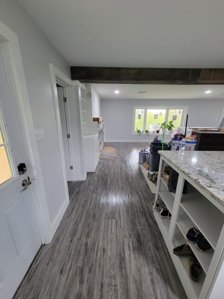 A hallway with wood-look flooring leads from a white door toward a sunlit room with a large window and a kitchen island.
