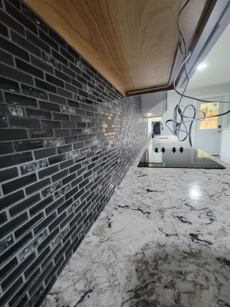 A high-angle view of a granite countertop and a dark, multi-textured glass brick tile backsplash under kitchen cabinets.