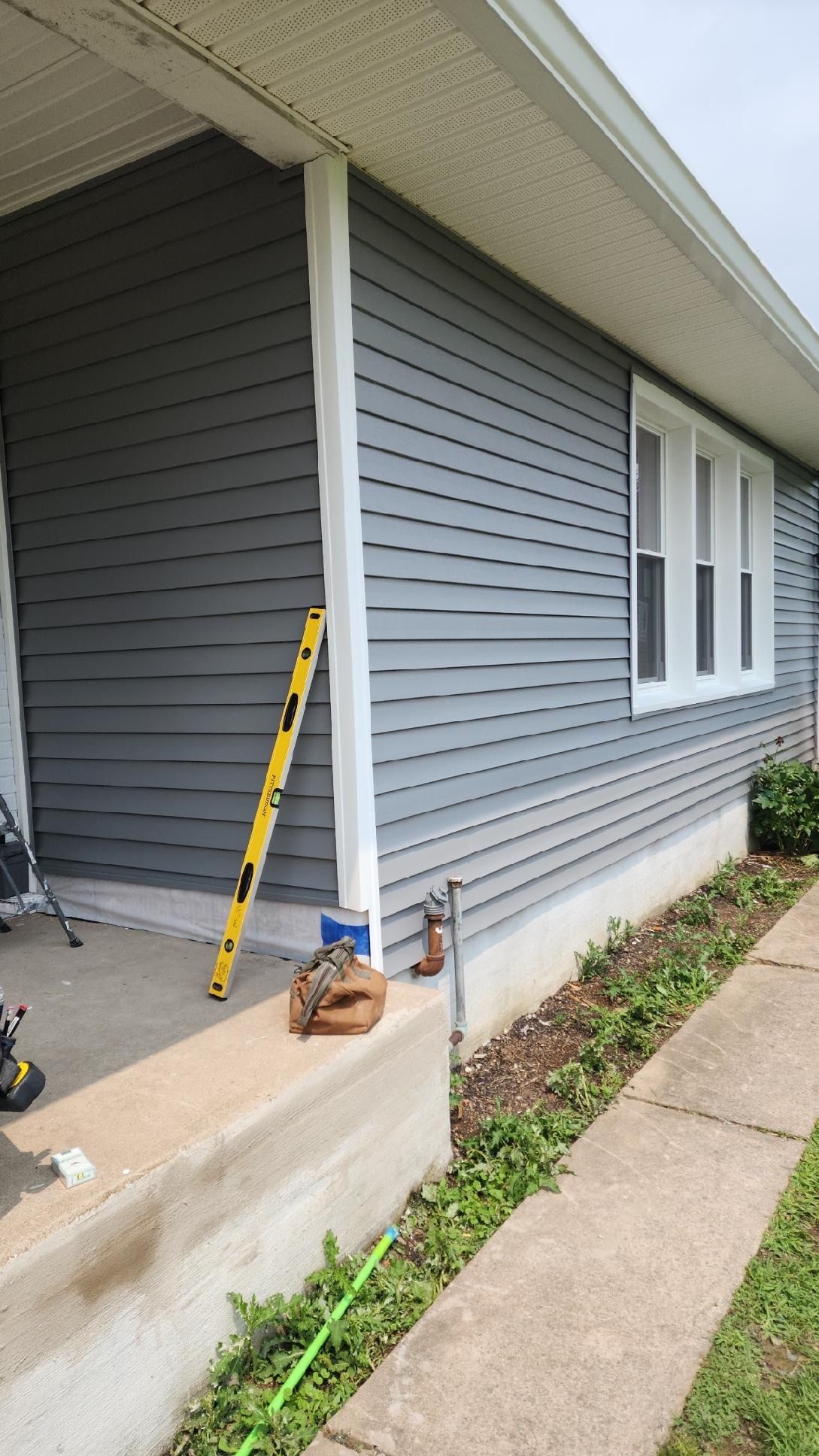 A side view of a house with gray vinyl siding, a white corner post, a yellow level leaning against the wall, and a porch.