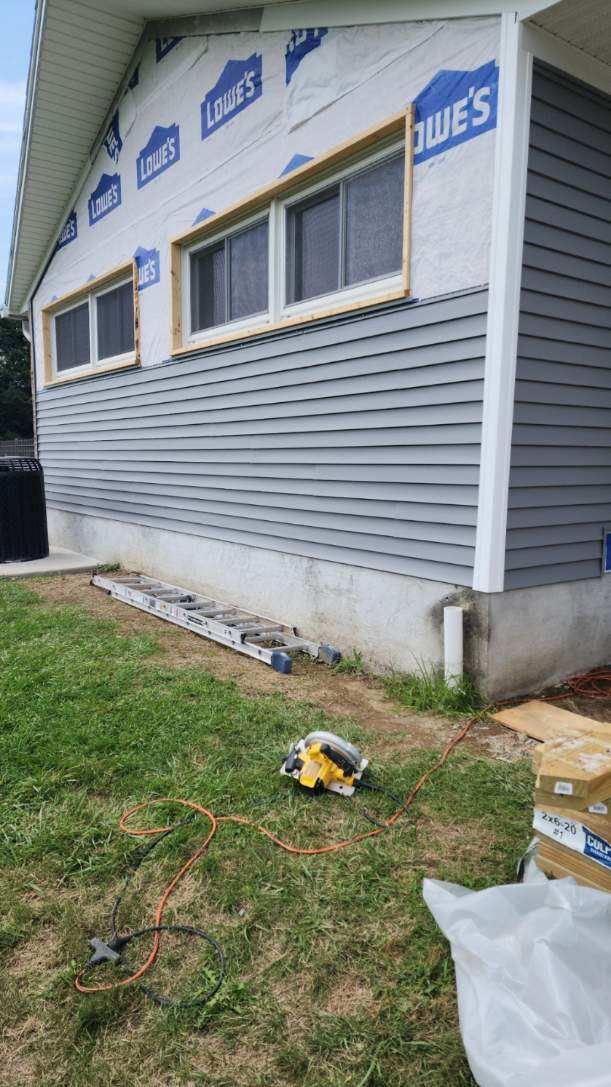 Gray vinyl siding is being installed on the side of a house, which has exposed wood framing and windows.