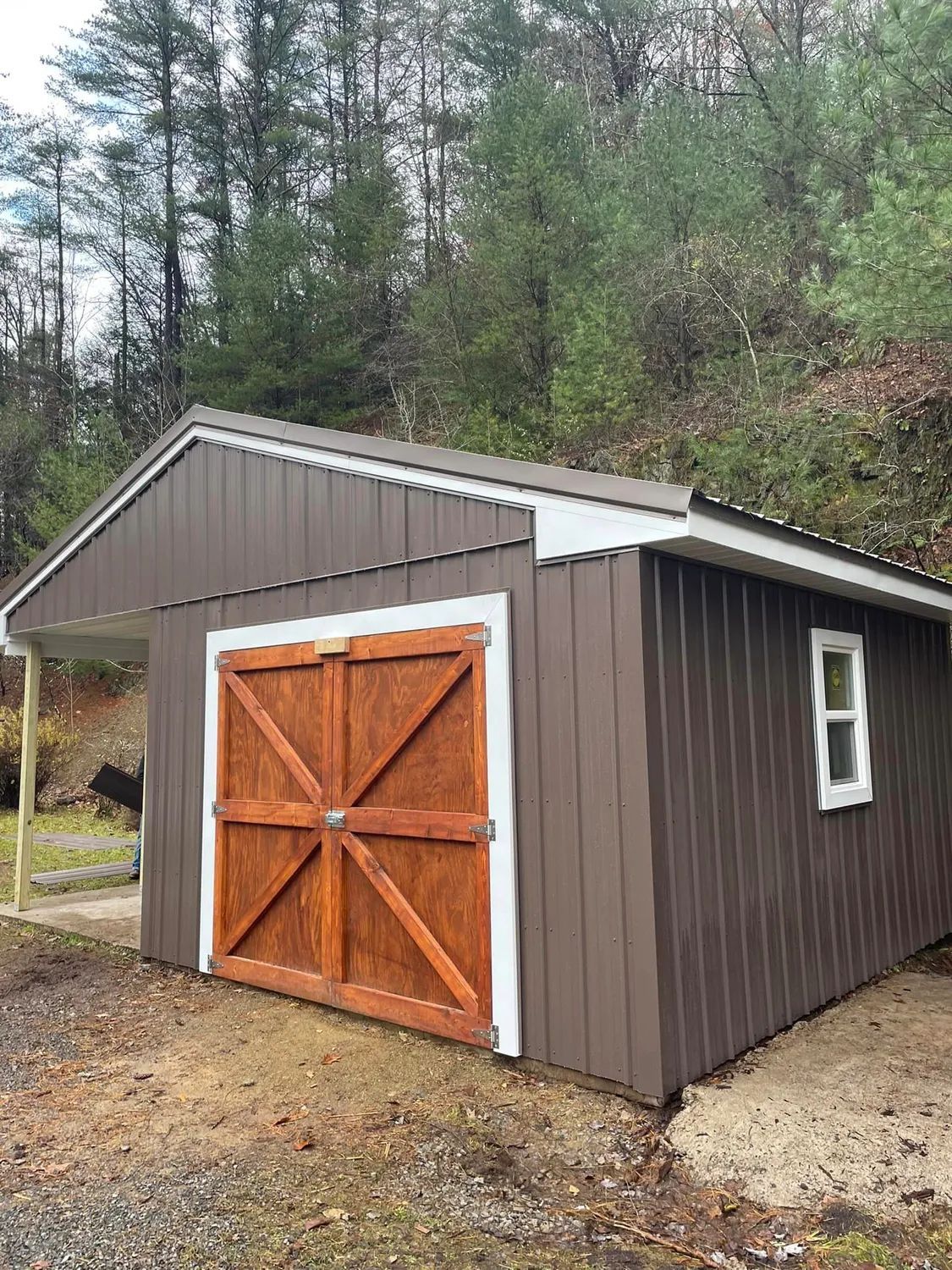 A brown wooden shed with double doors, a white-trimmed window, and an attached side porch sits in a wooded area.