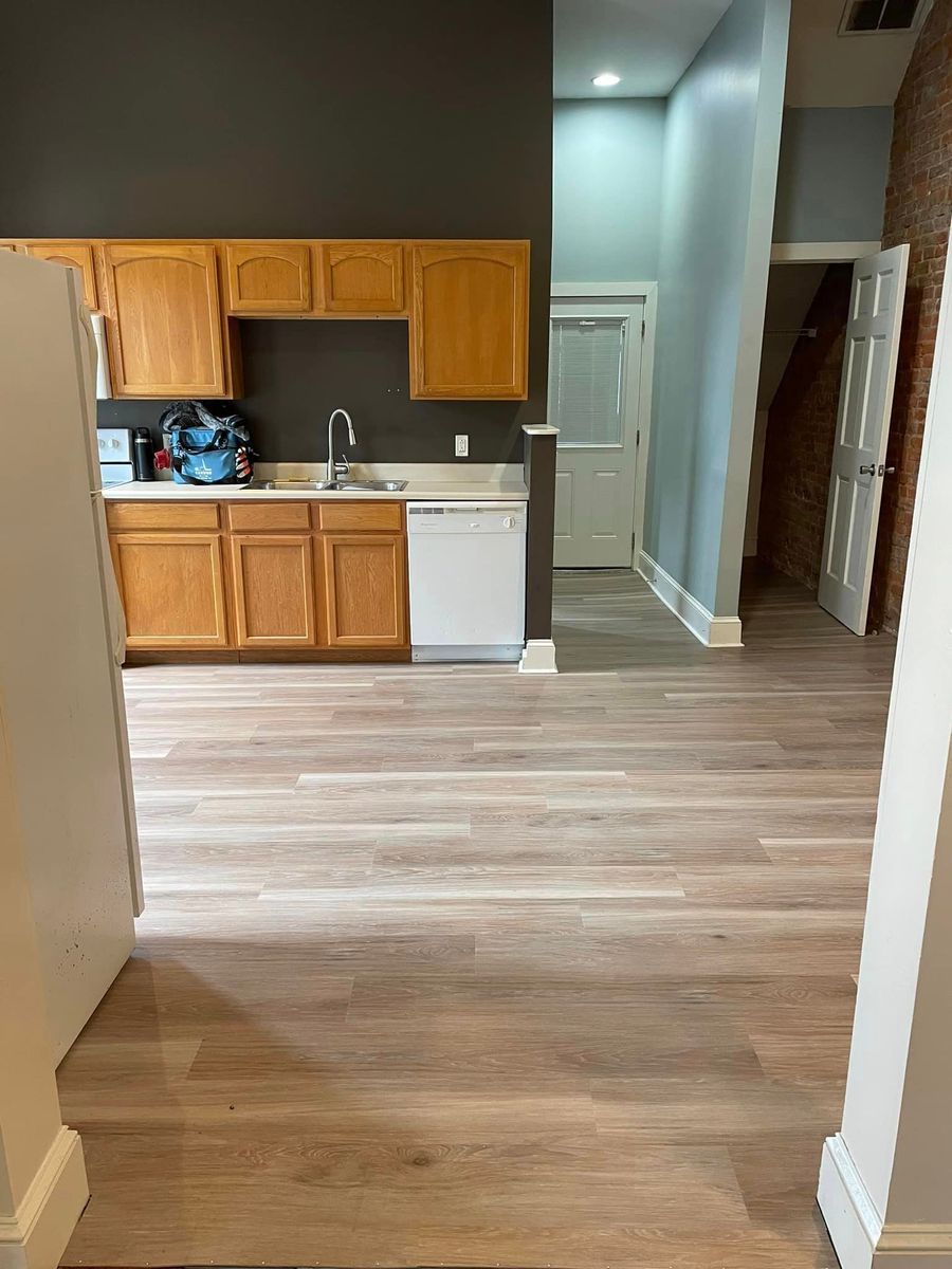 A kitchen with light wood-tone floors, medium-wood cabinets, a white dishwasher, and a doorway leading to another room.