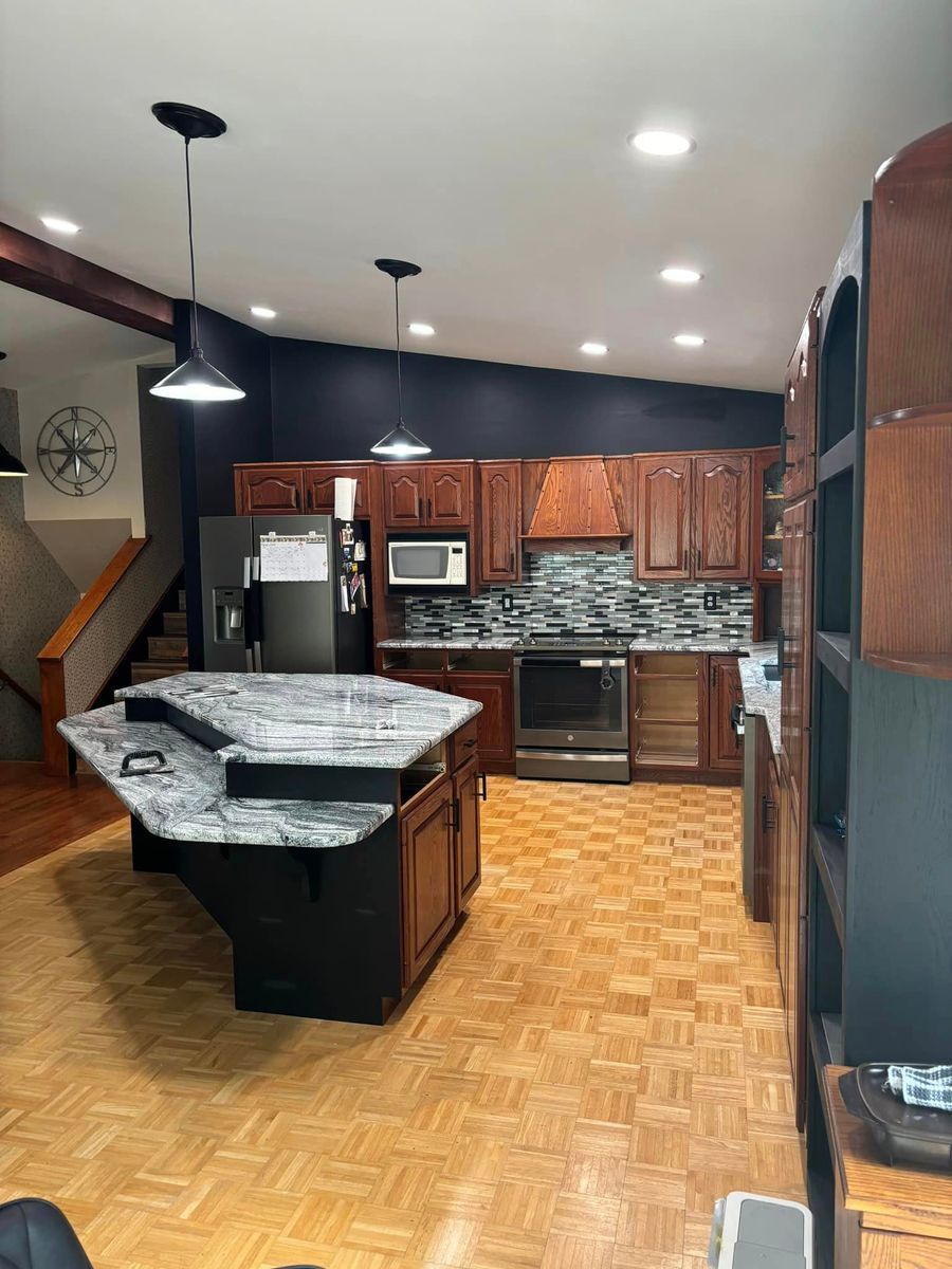 A kitchen with wood cabinets, a dark island with a granite countertop, stainless steel appliances, and parquet wood floors.