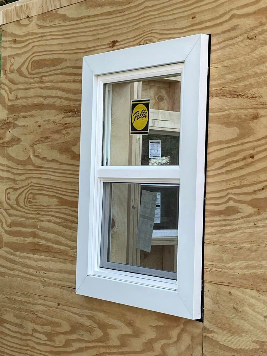 A white, single-hung window with a yellow sticker, installed in an unfinished plywood wall.