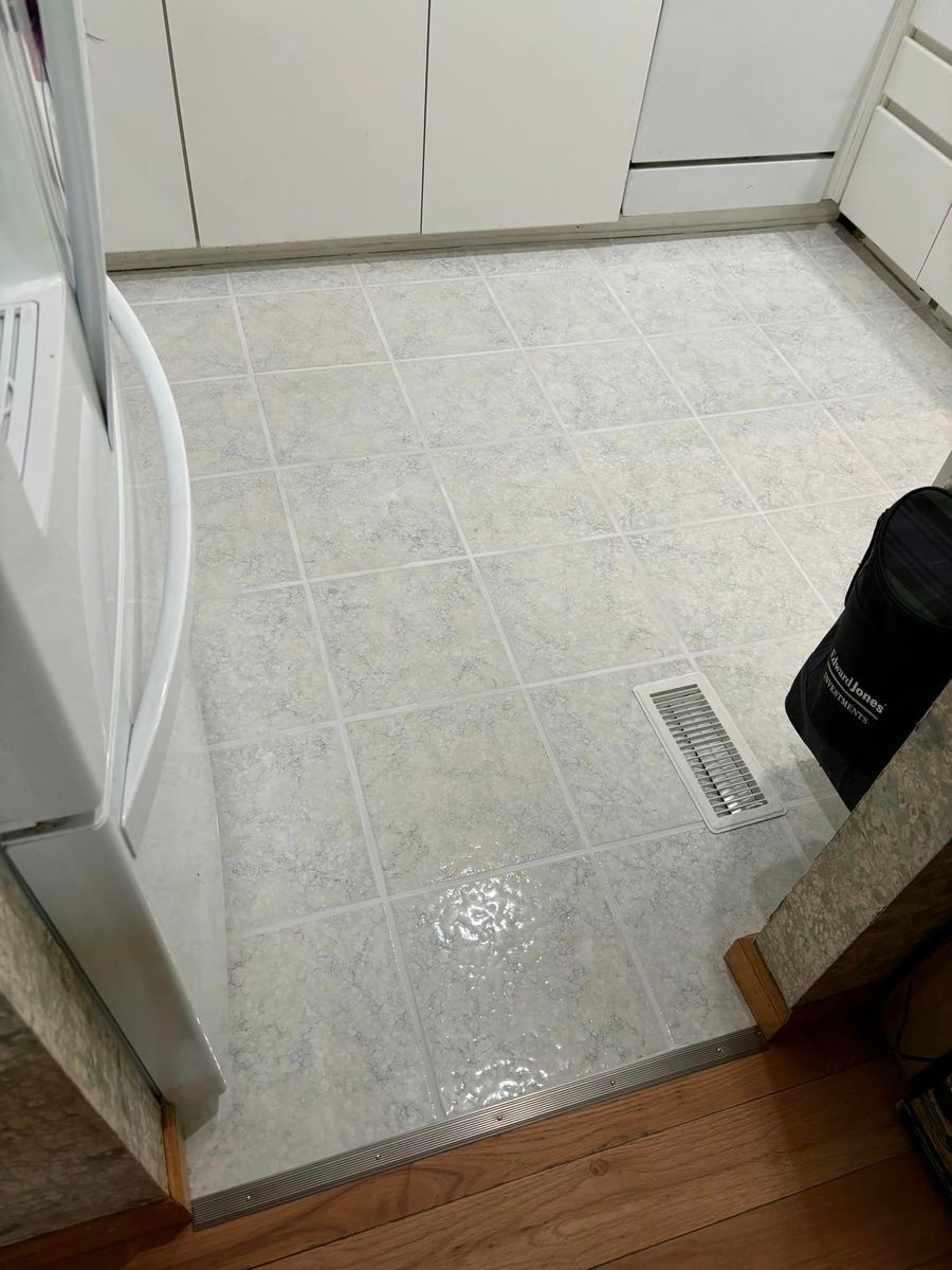 A kitchen floor with light, patterned square tiles, a white appliance on the left, and a floor air vent on the right.