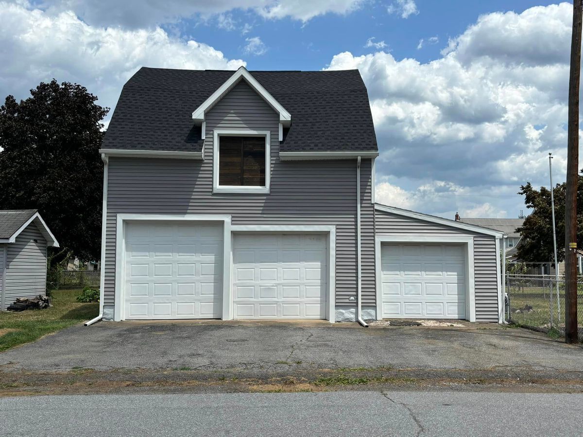 A two-story, gray-sided detached garage with three white garage doors and a gabled attic window under a cloudy blue sky.