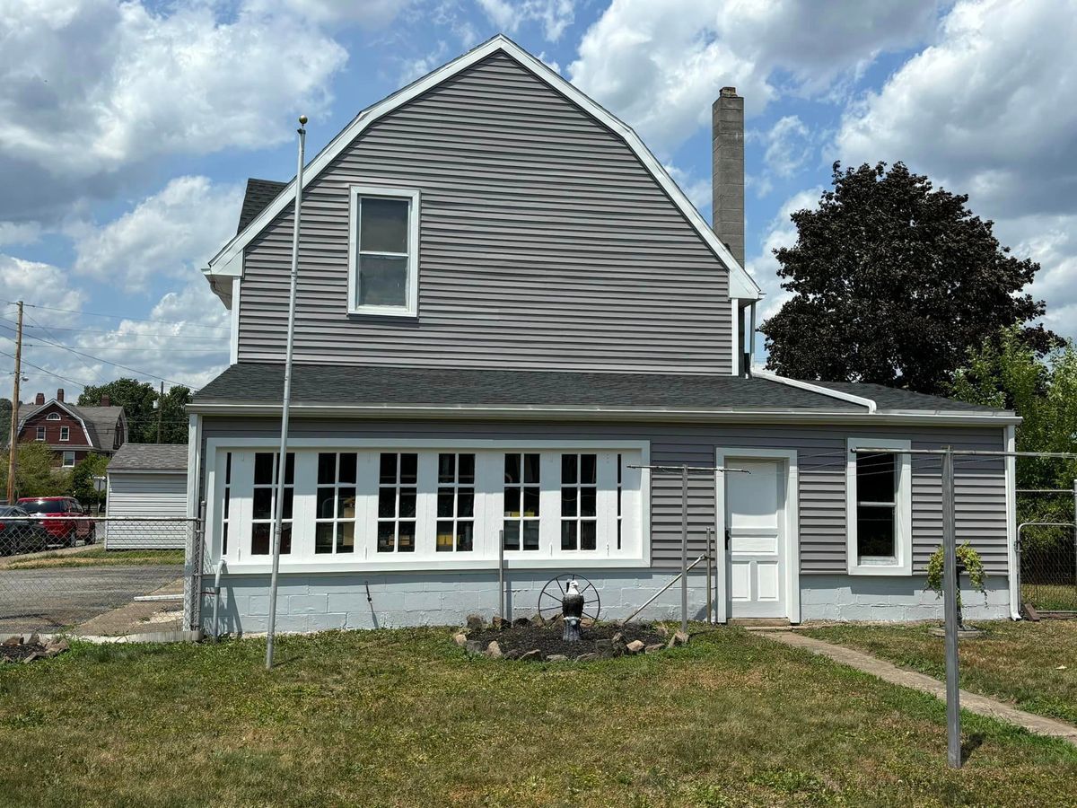 A two-story house with grey vinyl siding, a barn-style roof, a long multi-pane window, and a chimney against a blue sky.