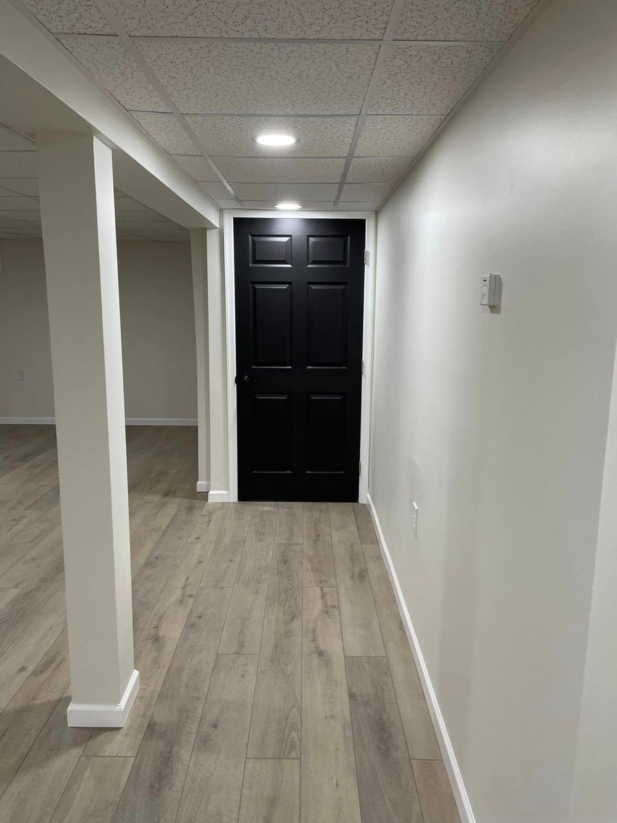 A hallway with wood-look flooring, light-colored walls, a drop ceiling with lights, and a black six-panel door at the end.
