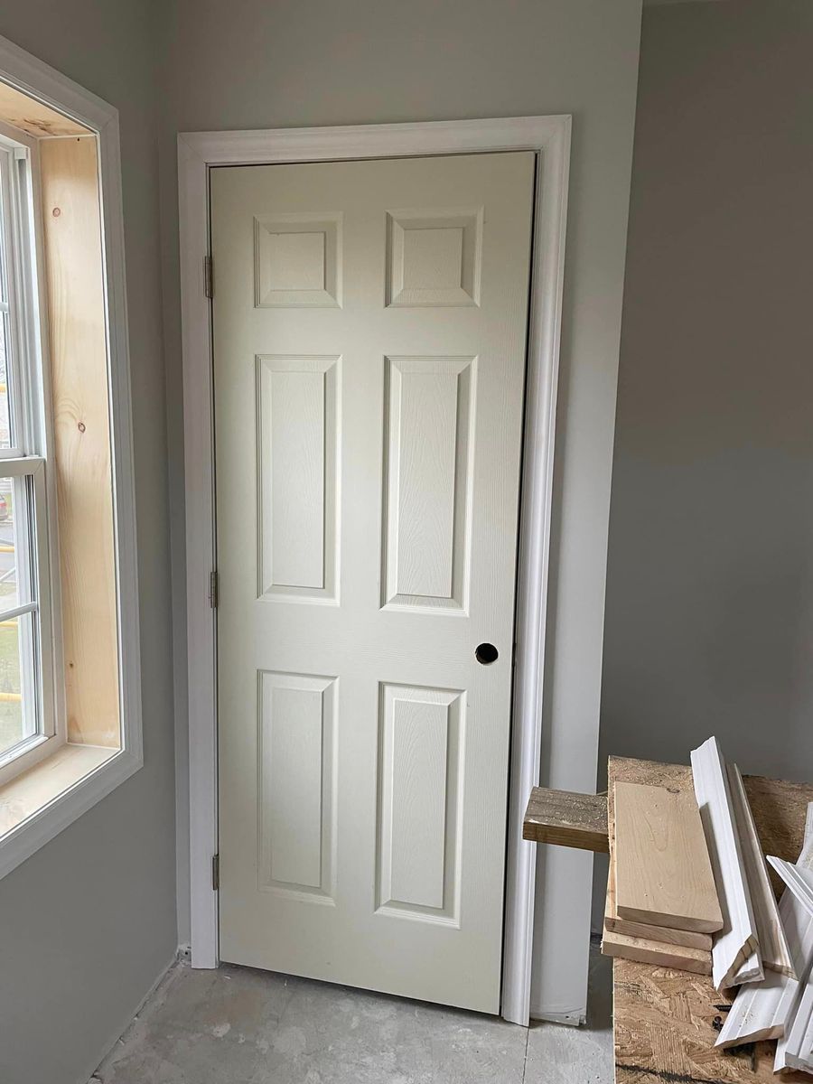 An interior, off-white, six-panel door with a dark knob installed in a white frame against gray walls near construction.