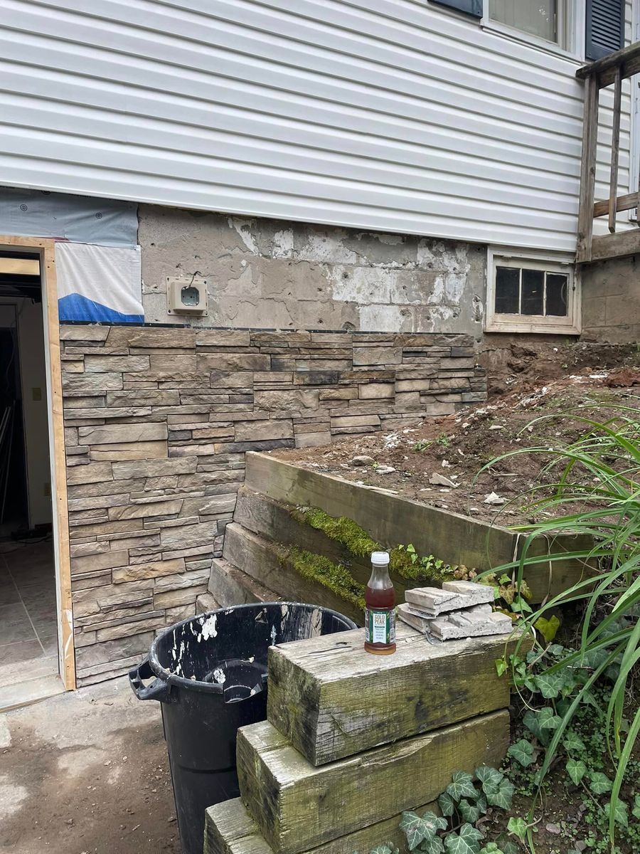 A partially completed exterior renovation with stone veneer on a foundation wall, near a retaining wall and trash can.