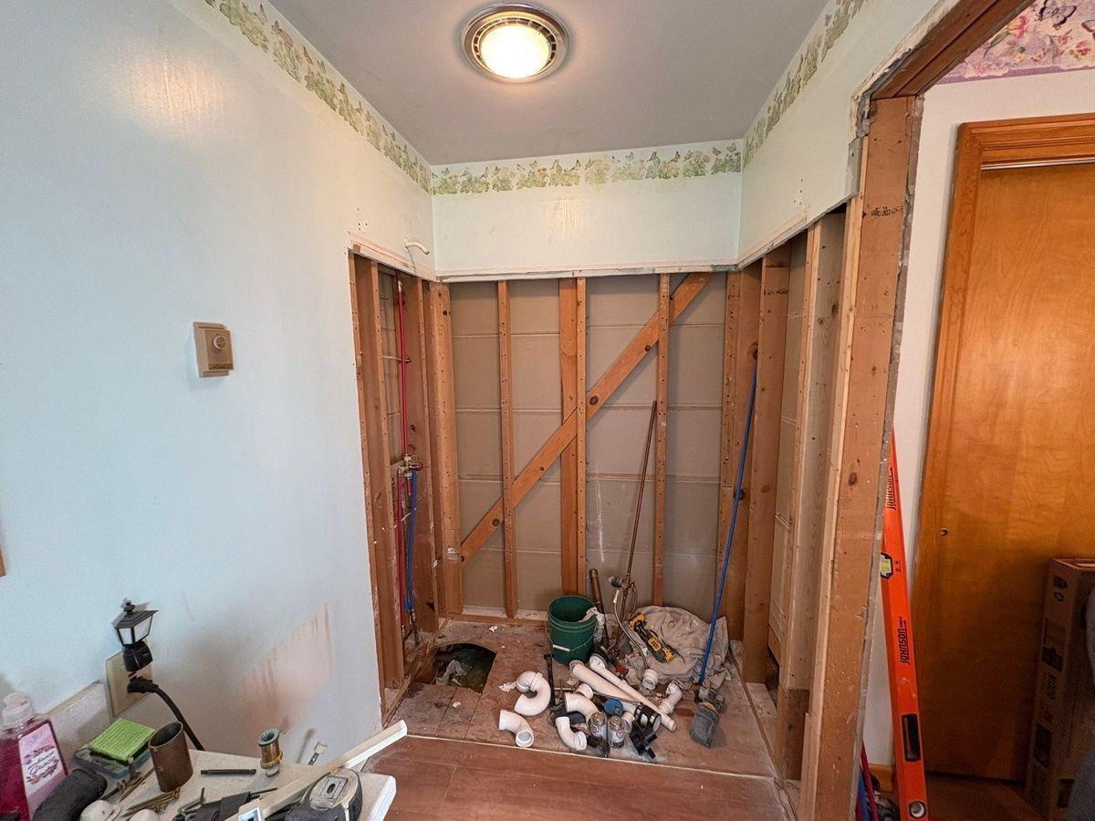 A construction scene showing an unfinished bathroom alcove with exposed wooden studs, plumbing pipes, and tools.