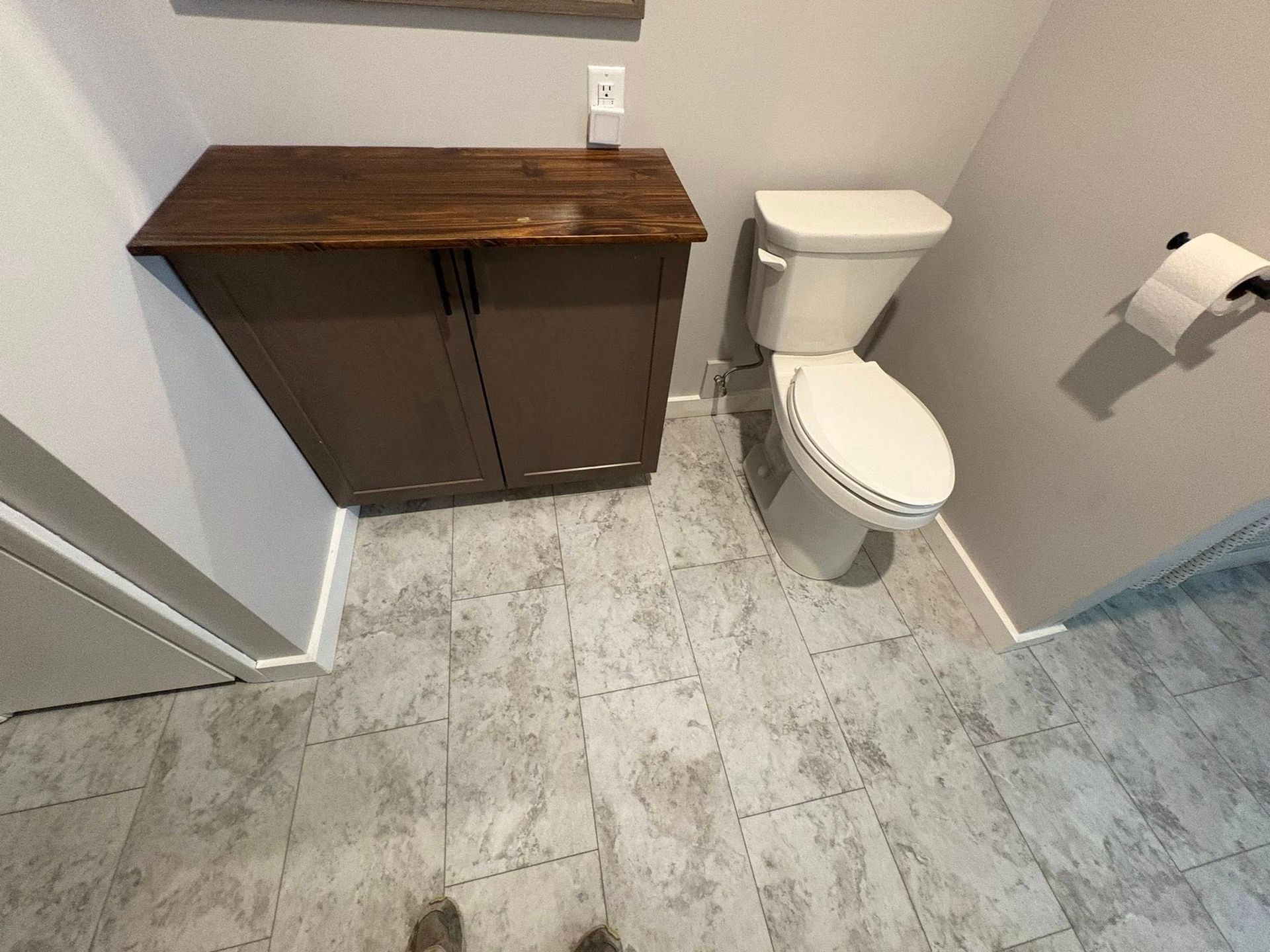A small brown cabinet with a dark wood top sits next to a white toilet in a bathroom with light gray tiled floors.