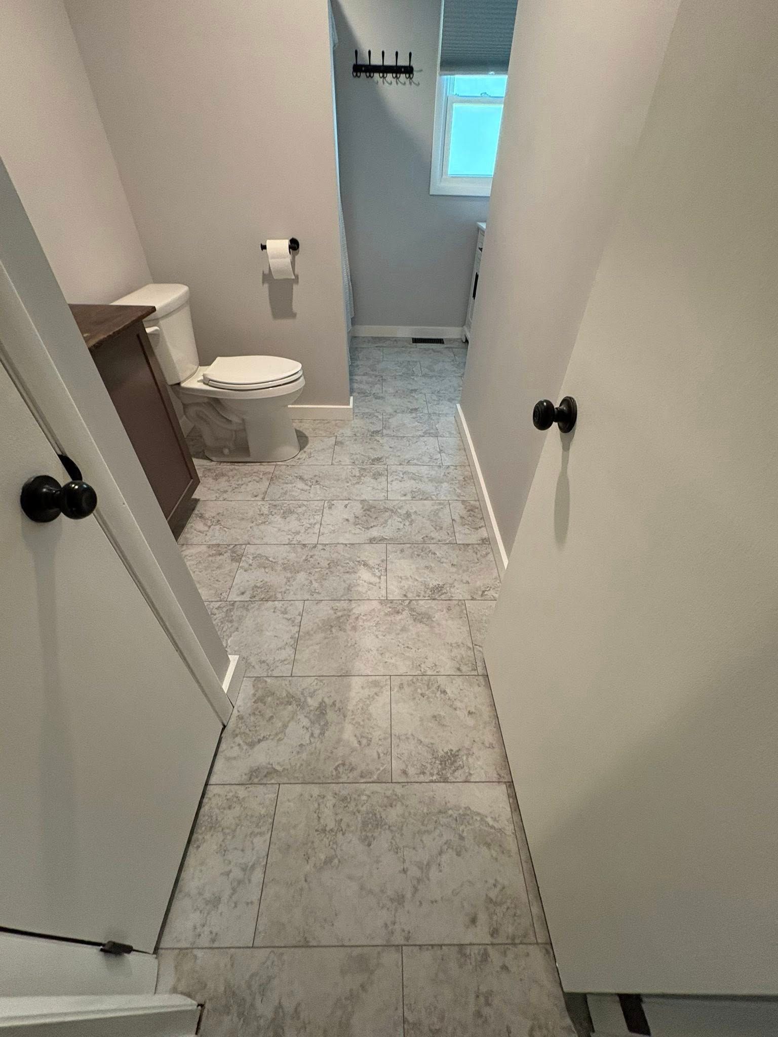A view through a doorway into a small bathroom with light gray tiled floors, a white toilet, and a vanity cabinet.