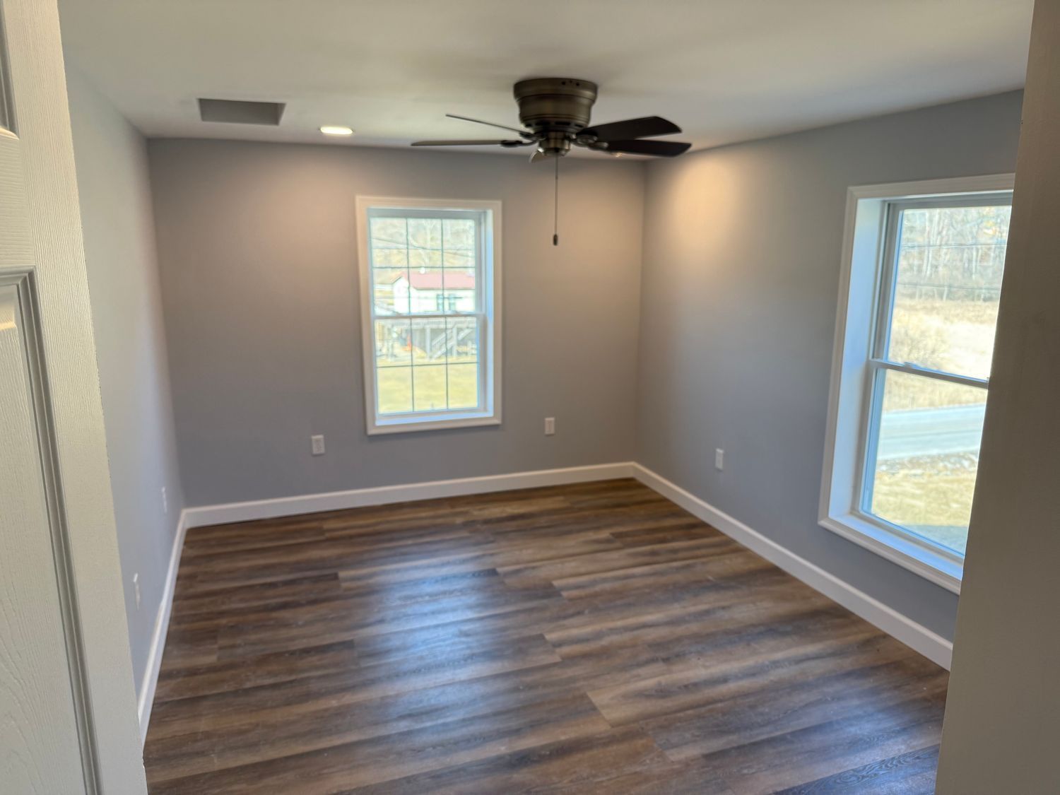 An empty room with gray walls, wood-look flooring, a ceiling fan, and two windows with outdoor views.