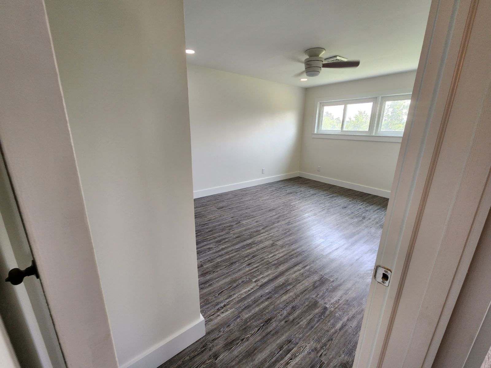 A view from a doorway into an empty room with white walls, gray wood-look flooring, and a ceiling fan.