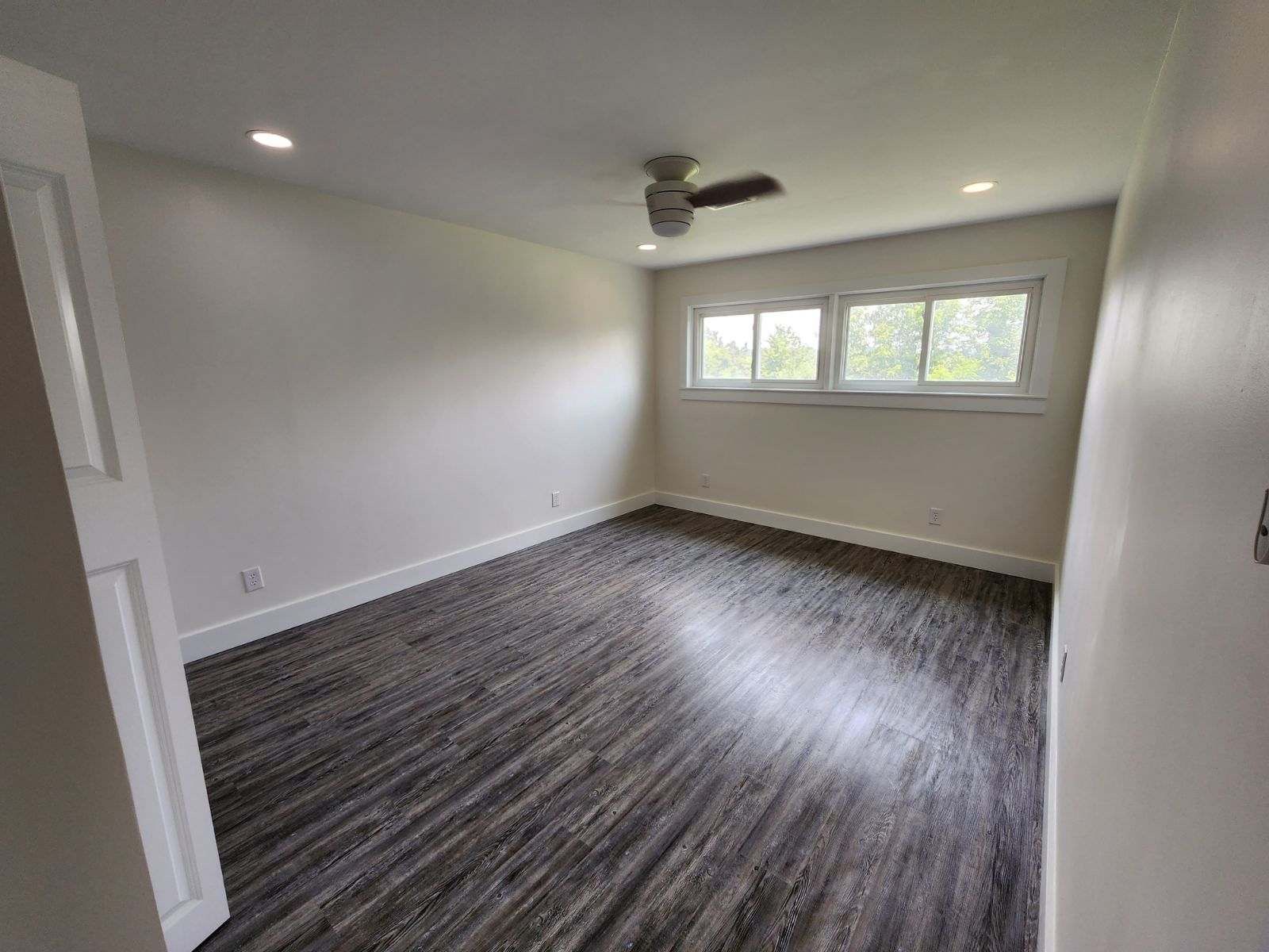 An empty bedroom with light-colored walls, wood-look flooring, a ceiling fan, and two windows facing the outdoors.