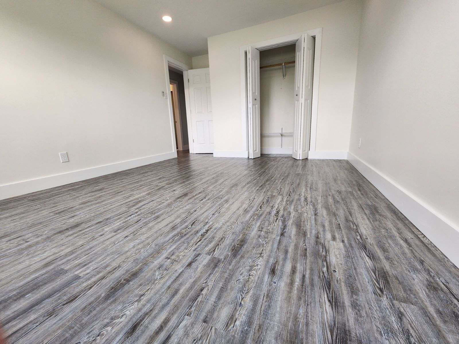 An empty room with grey wood-look flooring, white walls, a closet with white bifold doors, and a doorway to another room.