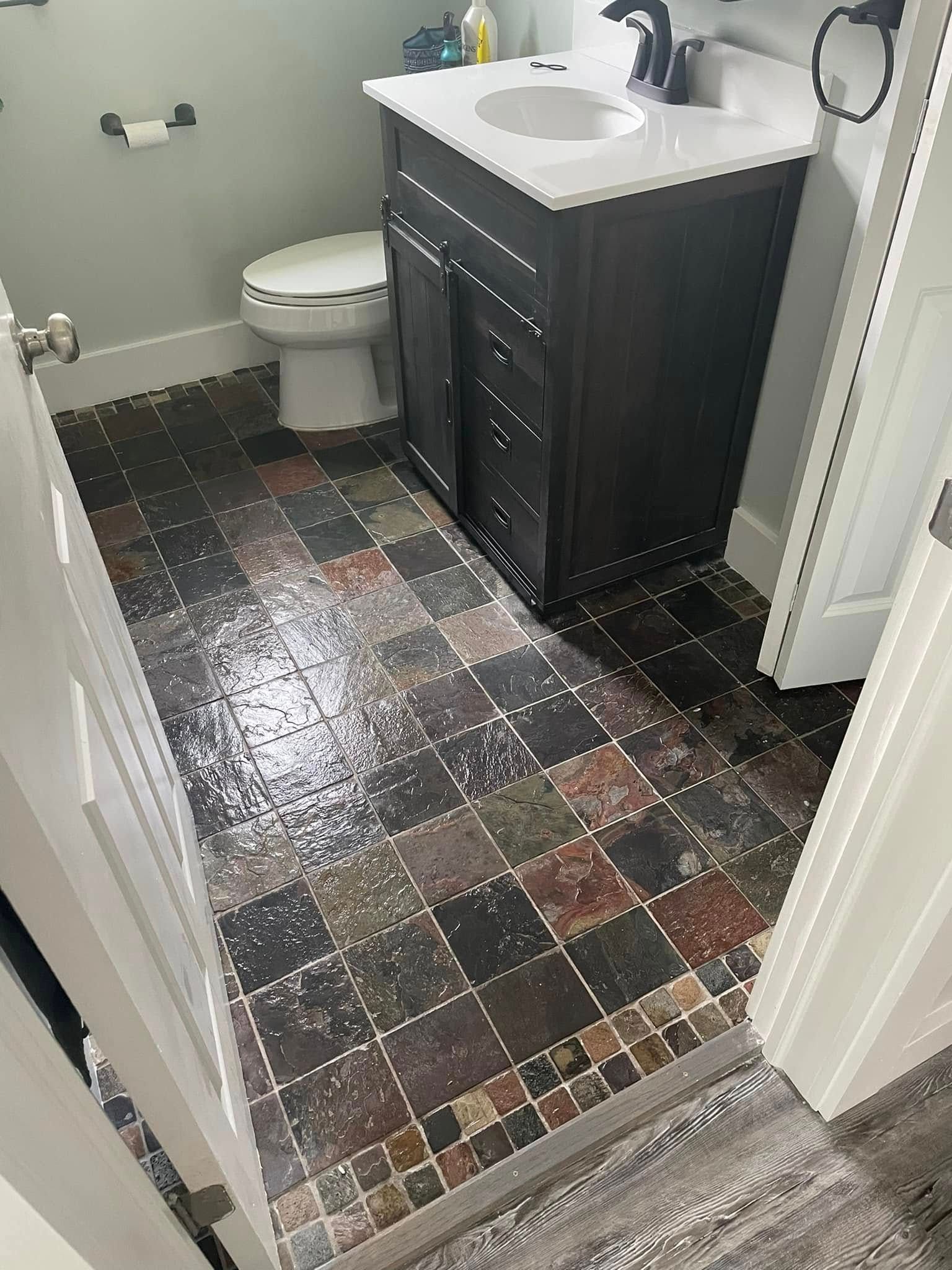 A small bathroom features a dark vanity with a white sink, a white toilet, and multi-colored, square tiled flooring.