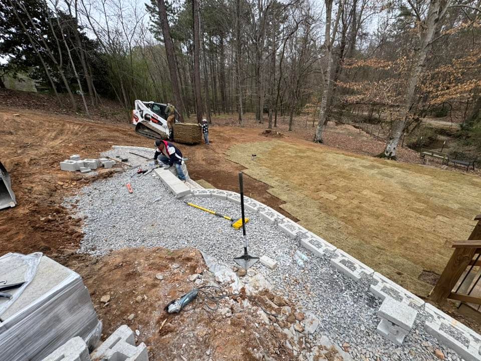 Construction site with retaining wall being built; gravel and cinder blocks visible. Person works with tools.