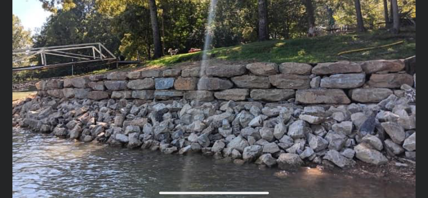 Stone retaining wall along a shoreline. Large rocks line the water's edge. Green grass and trees in background.
