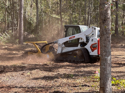 White Bobcat excavator and skid-steer loader digging in a wooded area, brown soil.
