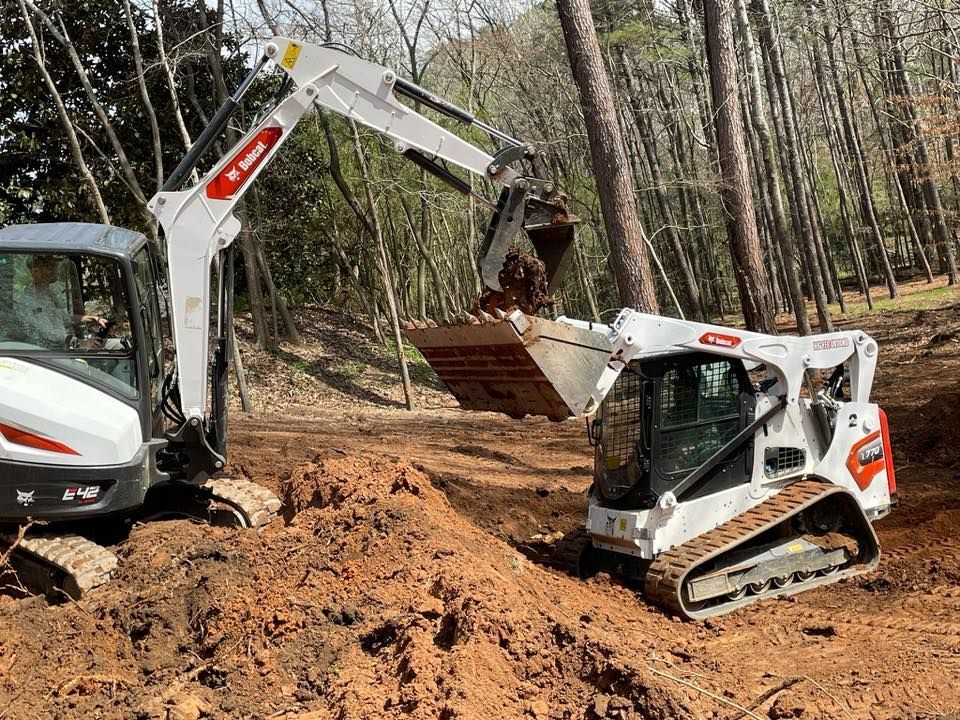 White Bobcat excavator and skid-steer loader digging in a wooded area, brown soil.