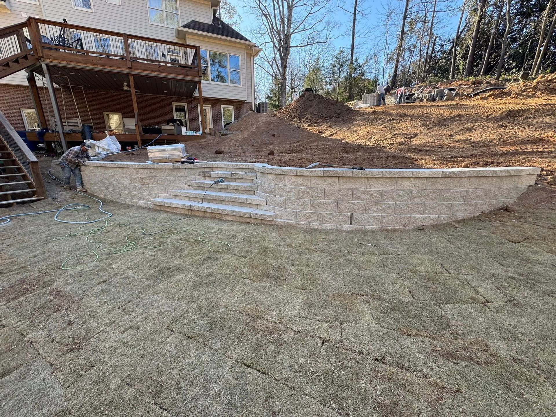 Stone retaining wall with steps, next to a house with a wooden deck and a sloped yard.