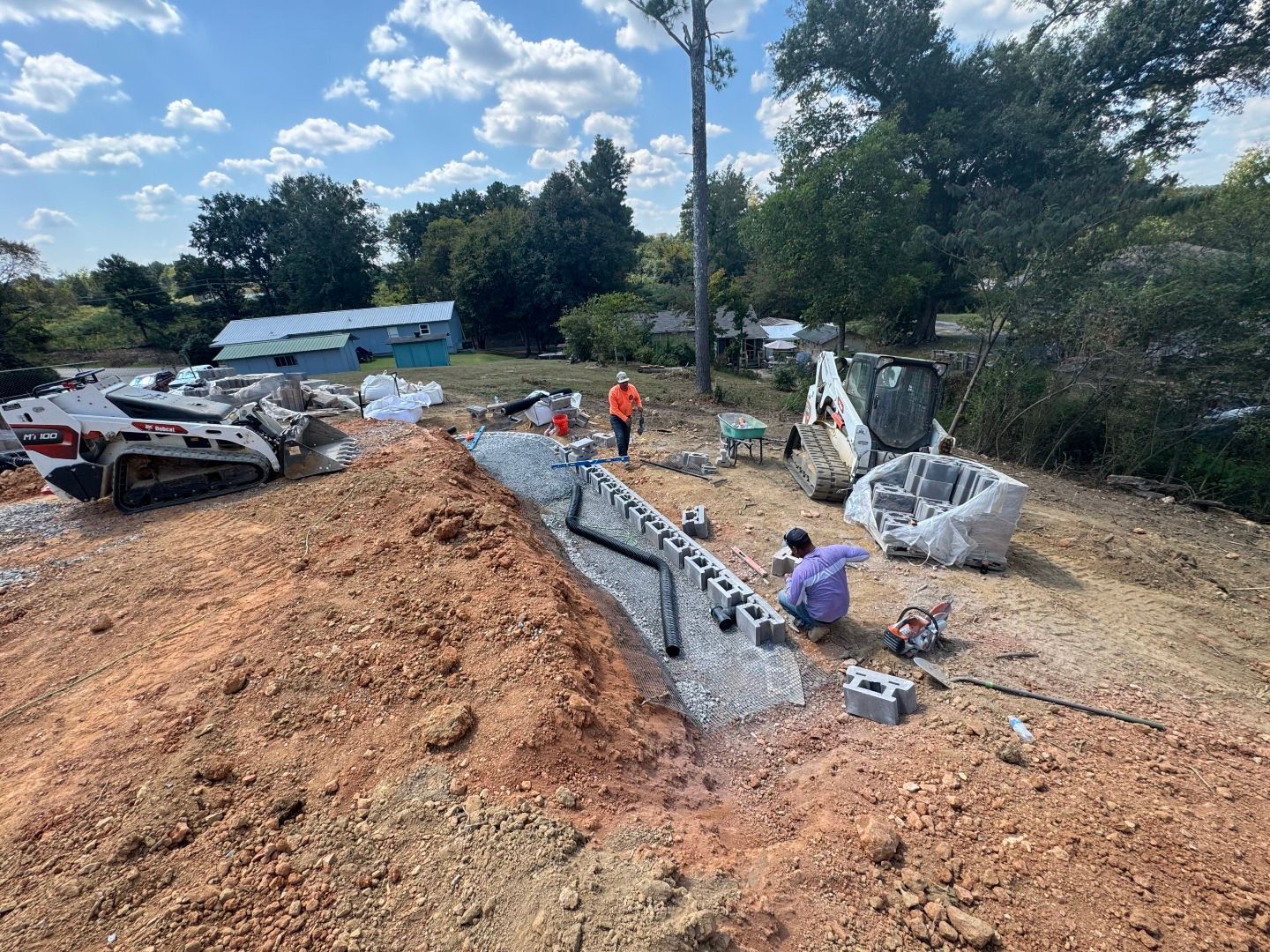 Construction site: Workers building retaining wall on a hillside. Red dirt, machinery, and trees in background.
