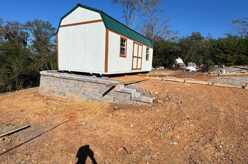 White shed with green roof on a retaining wall, with steps down to a dirt ground.