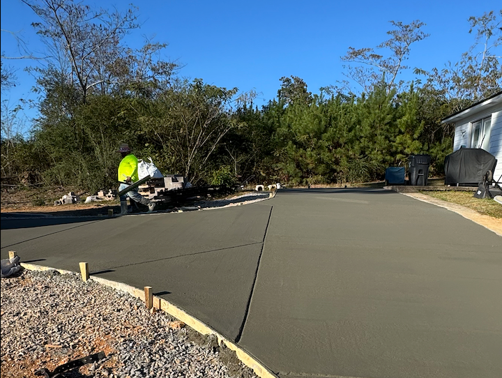Freshly poured concrete driveway with a worker, surrounded by trees and a house.