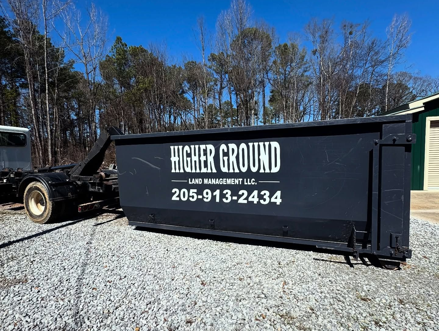 Black dumpster with Higher Ground LLC logo, phone number 205-913-2434, and a truck bed. Outdoors on gravel with trees.